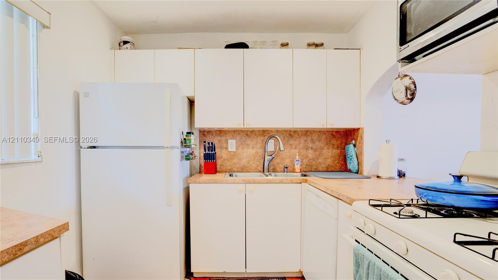 Kitchen featuring white cabinetry, solid-surface countertops, dual-basin stainless steel sink with high-arc faucet, and white appliances including a built-in microwave and gas range
