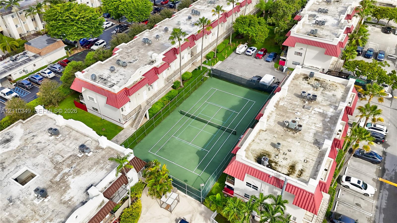 Aerial perspective of a multi-unit property featuring a fenced tennis court, red metal roofing, and surrounding palm trees