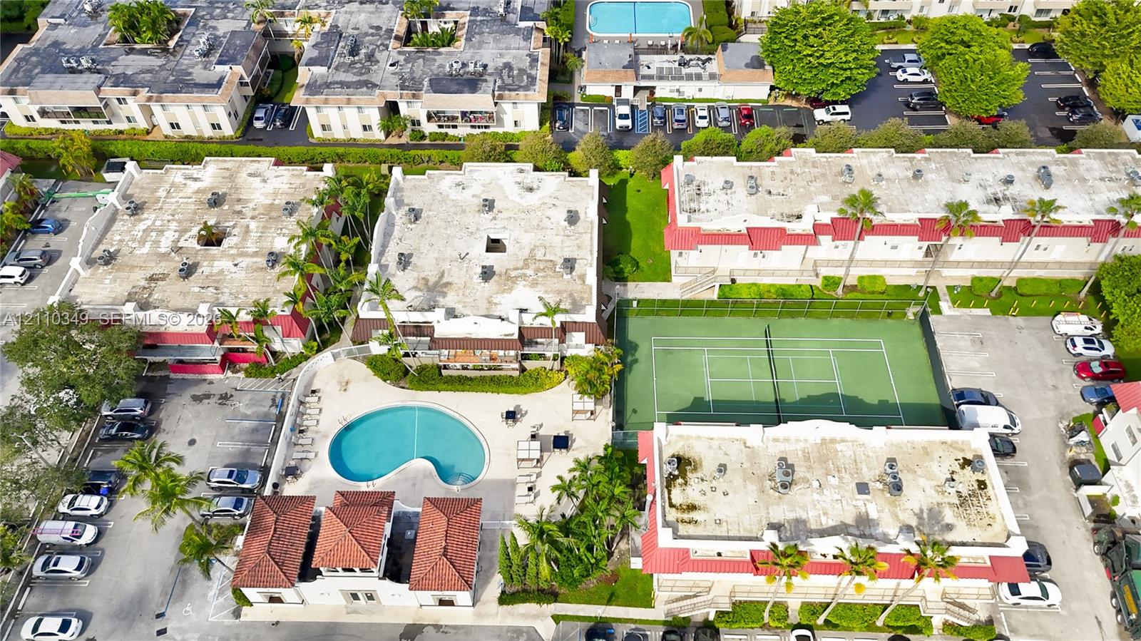 Aerial perspective of community grounds featuring a swimming pool, tennis court, tiled roof structures, and paved parking areas