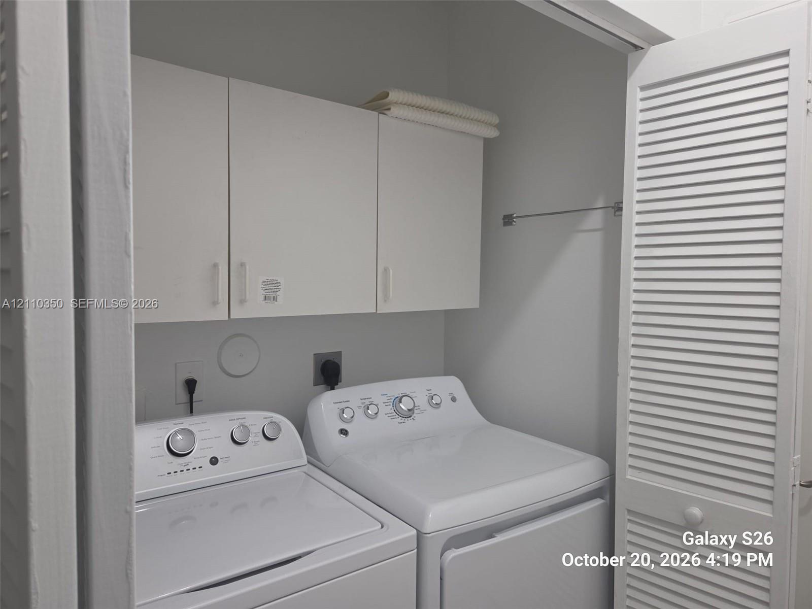 Laundry alcove featuring white louvered doors, upper wall-mounted cabinetry, and a horizontal metal hanging rod