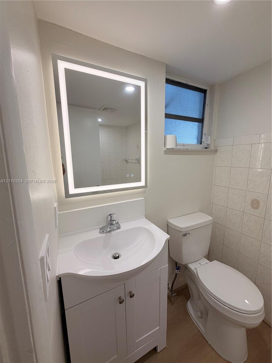 White vanity cabinet with an integrated basin, wall-mounted rectangular LED mirror, two-piece toilet, and tile wall detailing