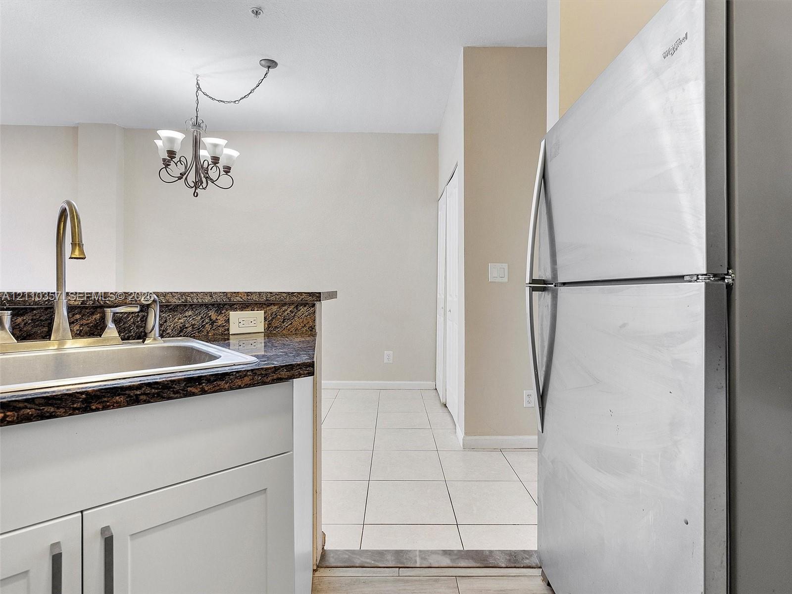 Kitchen featuring a stainless steel refrigerator, stone-surfaced countertop, stainless steel sink with high-arc faucet, white cabinetry, and multi-light chandelier