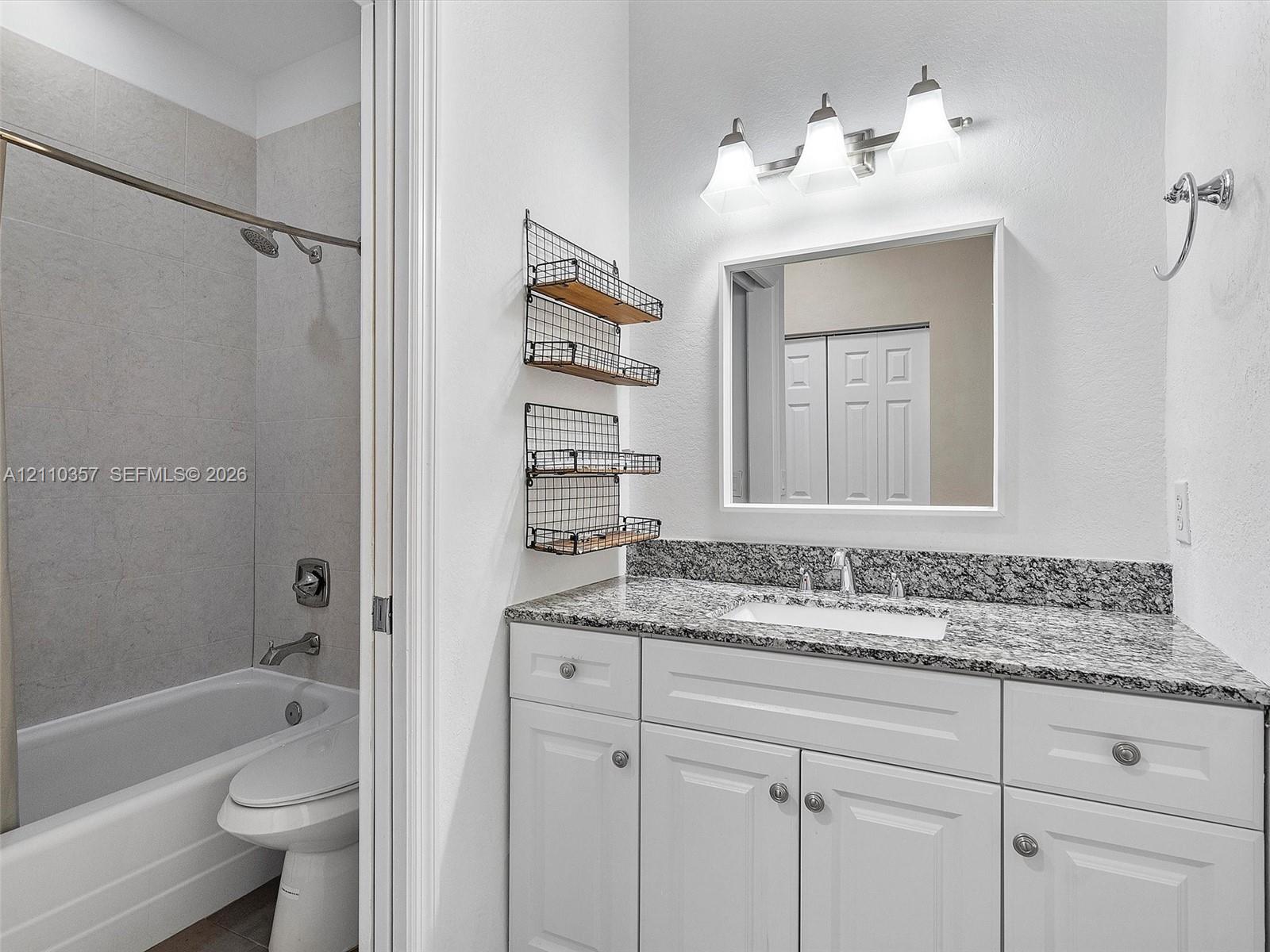 Bathroom featuring a white vanity with stone-patterned countertop, rectangular undermount sink, three-light fixture, and tub-shower combination with full-height tile surround