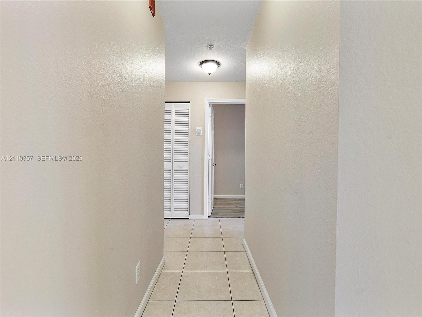 Interior hallway featuring neutral-toned walls, tile flooring, flush-mount ceiling lighting, and floor-to-ceiling louvered closet doors