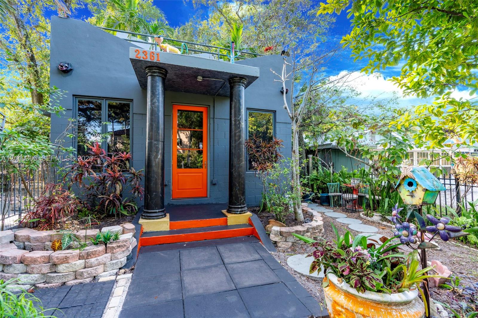 Exterior facade featuring structural support columns, a covered entryway, a flat-roof terrace with metal railing, and decorative stone-block garden retaining walls