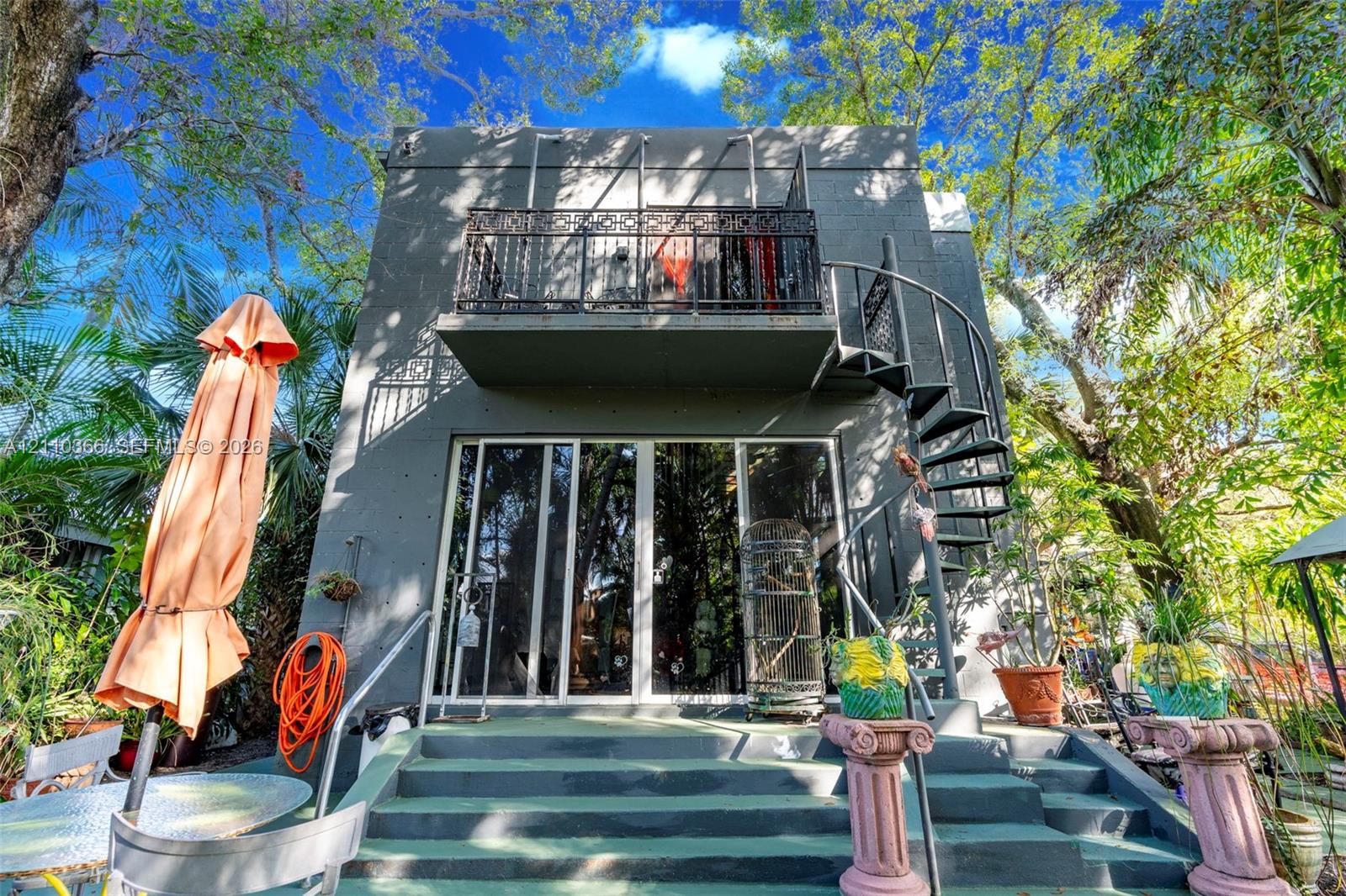 Multi-level exterior featuring a painted masonry facade, second-story balcony with decorative metal railing, exterior spiral staircase, and full-height glass sliding doors opening to a concrete staircase