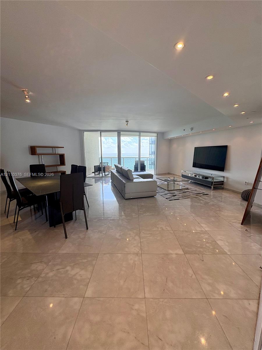 Open-concept living area featuring large-format tile flooring, recessed ceiling lighting, and floor-to-ceiling glass sliding doors leading to a balcony Open-concept living area featuring large-format tile flooring, recessed ceiling lighting, and floor-to-ceiling glass sliding doors leading to a balcony