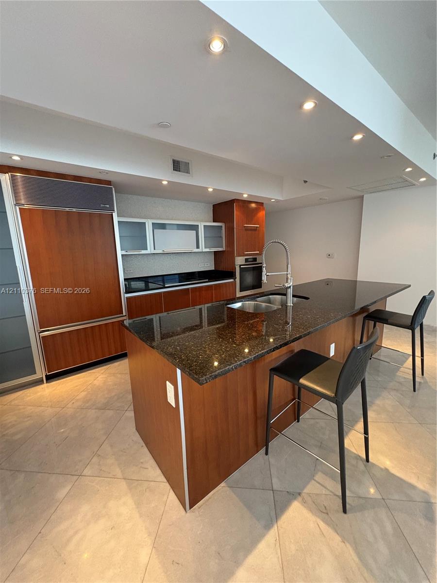 Kitchen featuring wood cabinetry, integrated refrigerator, dark stone countertops, central island with double-basin sink, and tile flooring Kitchen featuring wood cabinetry, integrated refrigerator, dark stone countertops, central island with double-basin sink, and tile flooring