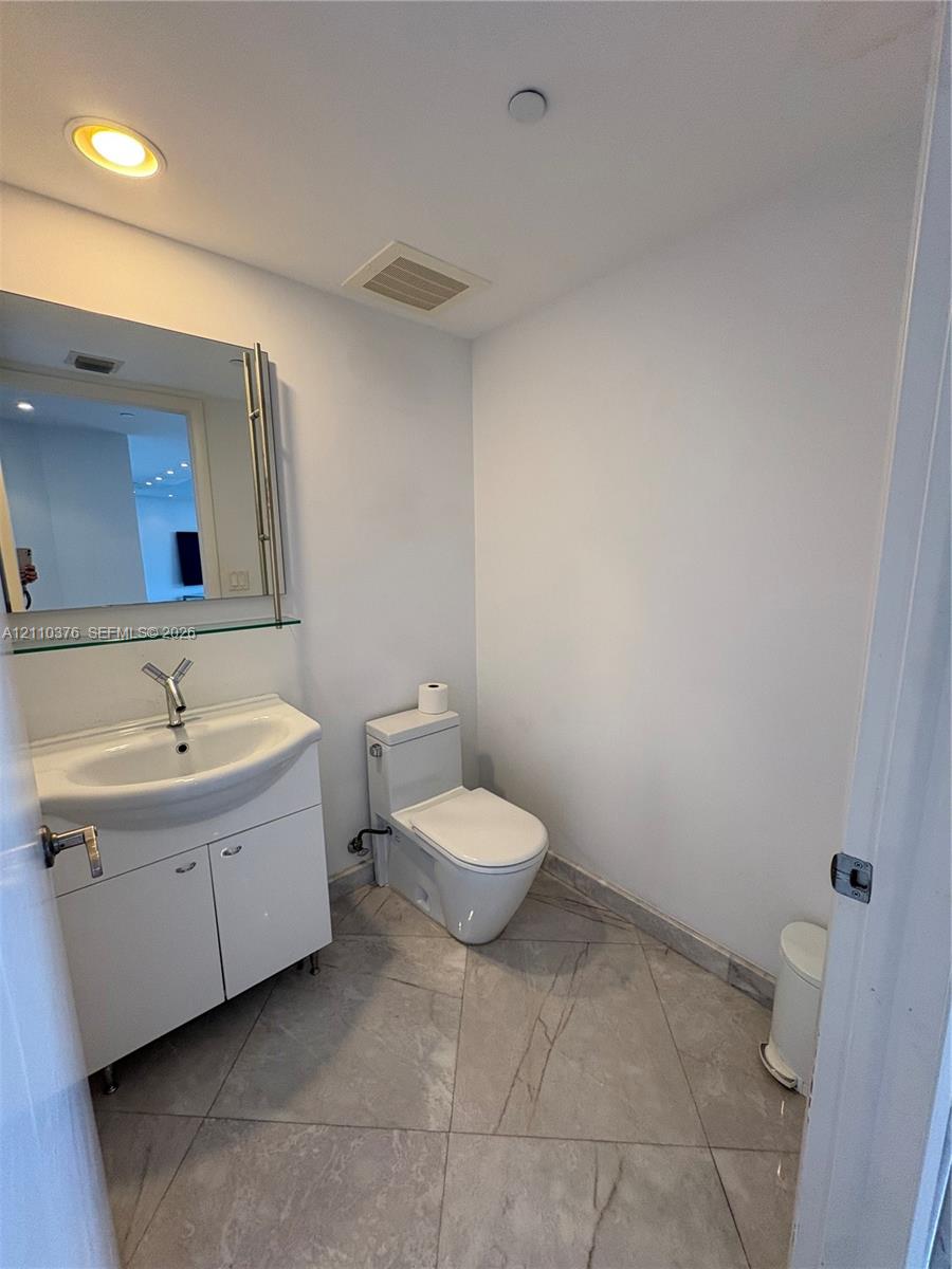 White cabinetry vanity with integrated sink, polished metal faucet, wall-mounted mirror with glass shelving, and large-format tile flooring