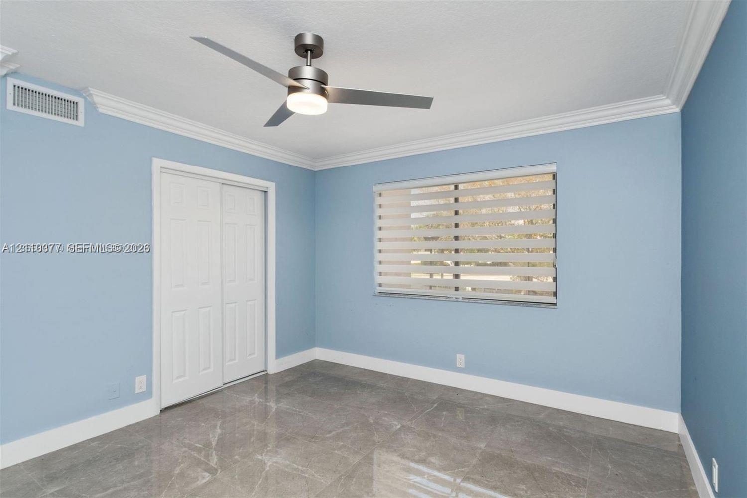 Large-format stone-finish tile flooring, matte metallic ceiling fan with integrated lighting, crown molding, and dual-panel closet doors