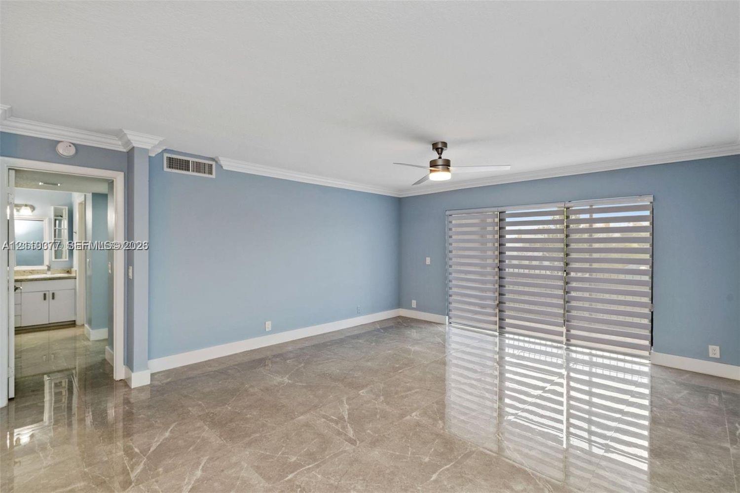 Open living area featuring large-format stone-finish tile flooring, crown molding, a ceiling-mounted light fixture with fan, and floor-to-ceiling sliding glass doors with horizontal blinds