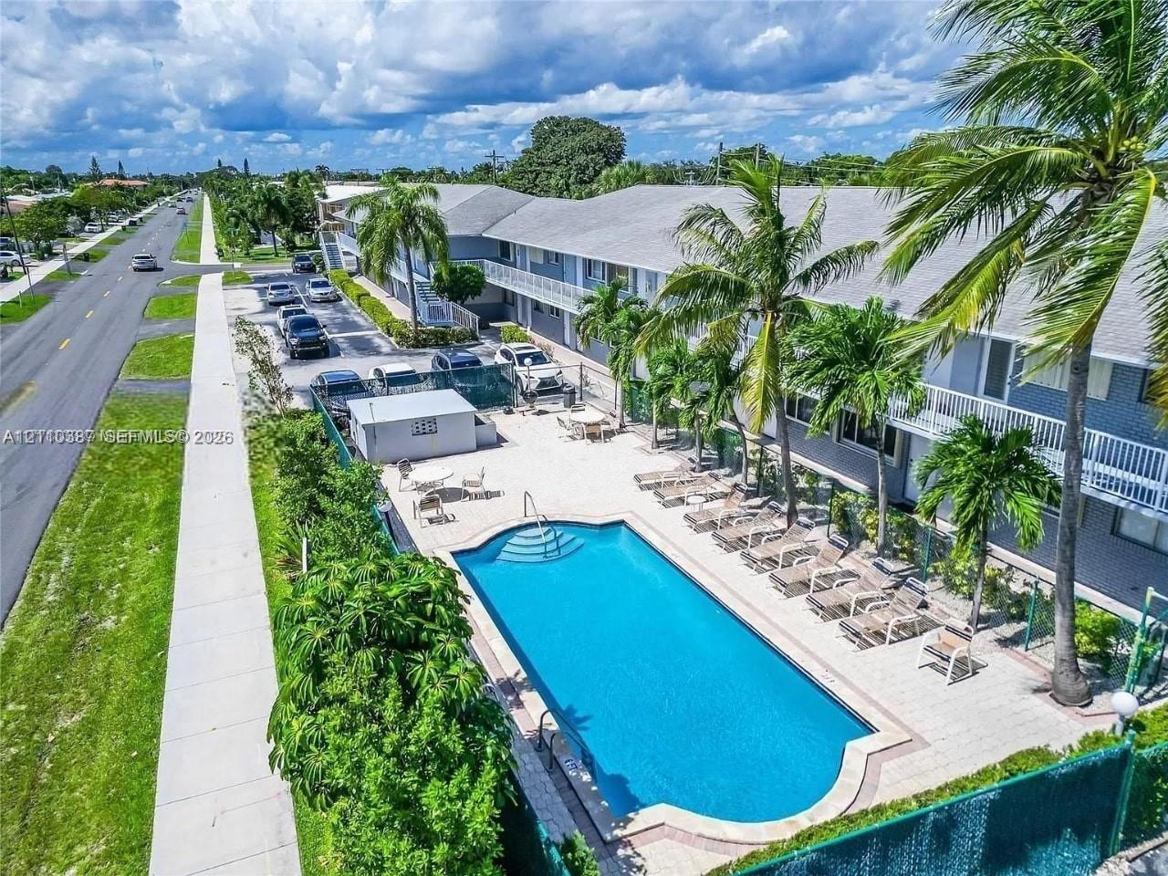 In-ground swimming pool with surround decking, mature palm trees, multi-story structure with white railings, and paved surface parking