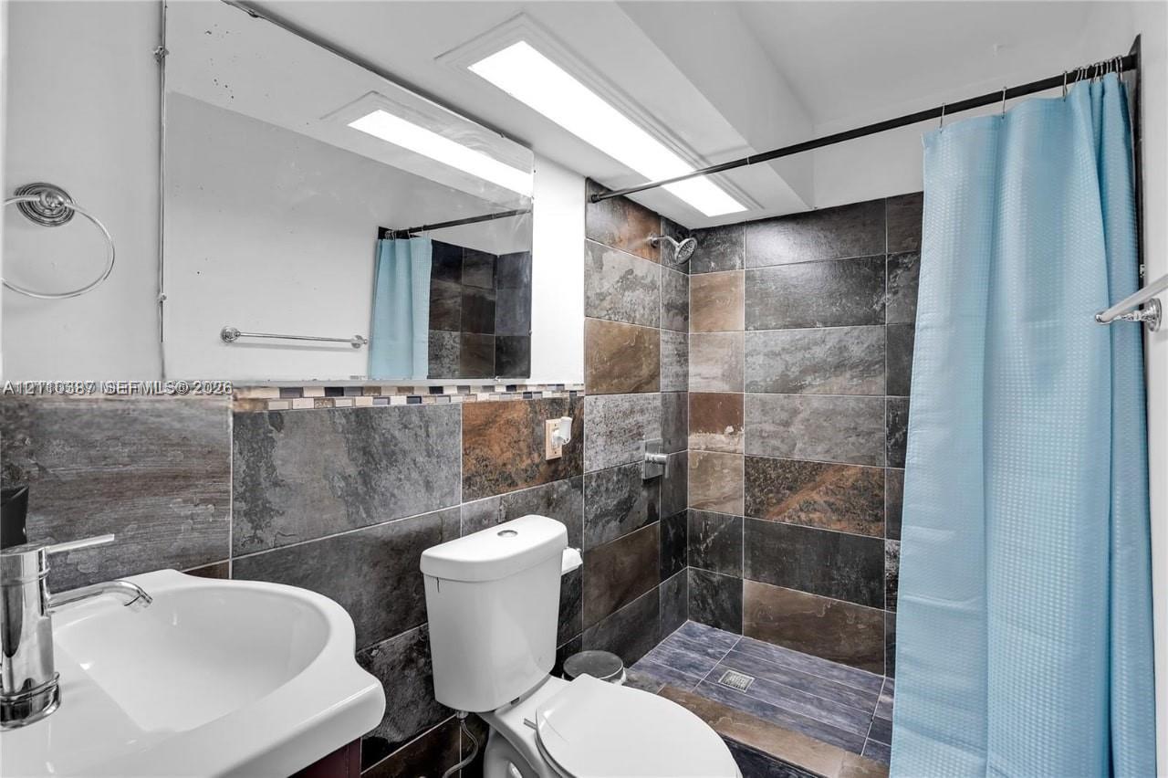 Bathroom featuring large-format multicolored stone wall tiling, a pedestal sink with a polished chrome faucet, a wall-mounted mirror, and a walk-in shower area with recessed lighting Bathroom featuring large-format multicolored stone wall tiling, a pedestal sink with a polished chrome faucet, a wall-mounted mirror, and a walk-in shower area with recessed lighting
