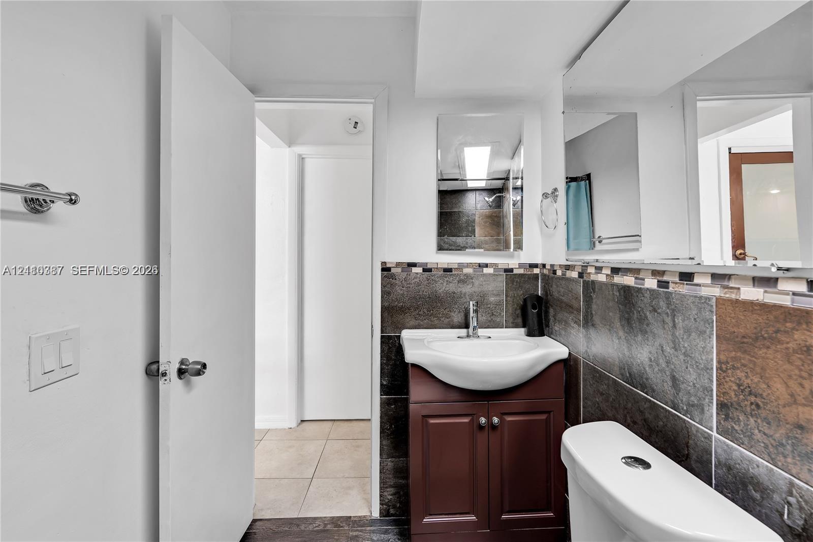 Bathroom featuring a dark wood-finish vanity with a ceramic basin, chrome faucet, expansive wall-to-wall mirror, and floor-to-ceiling tilework with mosaic decorative accents Bathroom featuring a dark wood-finish vanity with a ceramic basin, chrome faucet, expansive wall-to-wall mirror, and floor-to-ceiling tilework with mosaic decorative accents