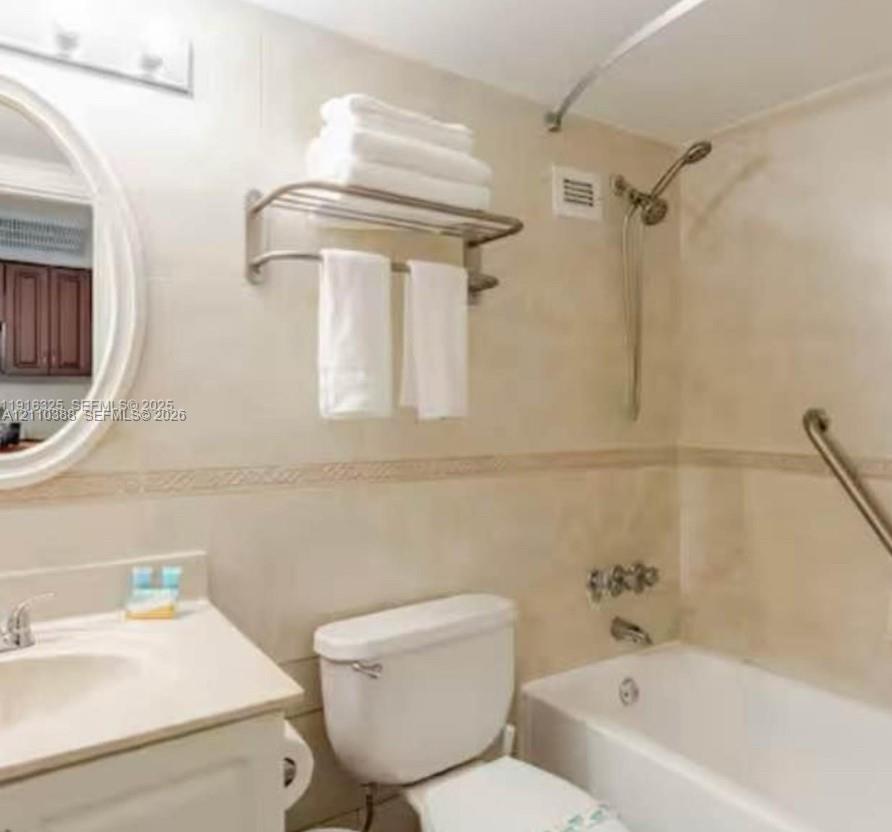 Bathroom featuring floor-to-ceiling tile, oval wall mirror, integrated vanity countertop with sink, and a combination tub and shower with metallic hardware Bathroom featuring floor-to-ceiling tile, oval wall mirror, integrated vanity countertop with sink, and a combination tub and shower with metallic hardware