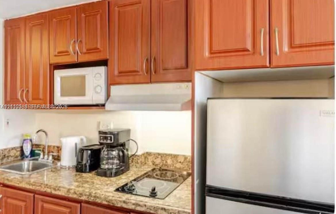 Kitchen featuring wood-toned cabinetry, stone-patterned countertops, stainless steel sink with a high-arch faucet, integrated stovetop, and built-in microwave Kitchen featuring wood-toned cabinetry, stone-patterned countertops, stainless steel sink with a high-arch faucet, integrated stovetop, and built-in microwave