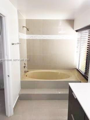 Built-in soaking tub with a stepped tile surround, wall-mounted shower fixture, decorative mosaic tile inlay, and horizontal window blinds