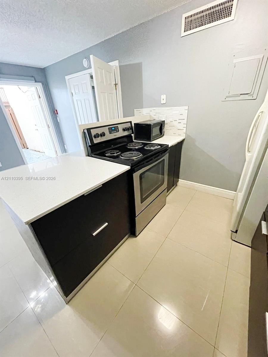 Kitchen featuring dark cabinetry, solid-surface countertops, tile backsplash, stainless steel range, and large-format tile flooring Kitchen featuring dark cabinetry, solid-surface countertops, tile backsplash, stainless steel range, and large-format tile flooring