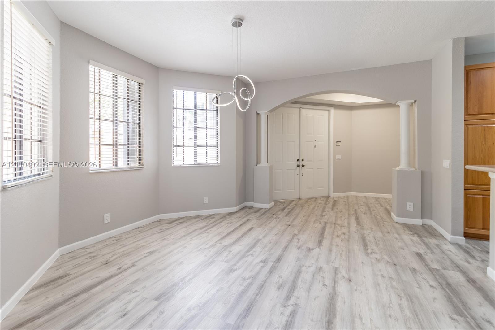 Wood-finish flooring, arched entryway with architectural columns, contemporary geometric pendant lighting, and multiple windows with horizontal blinds