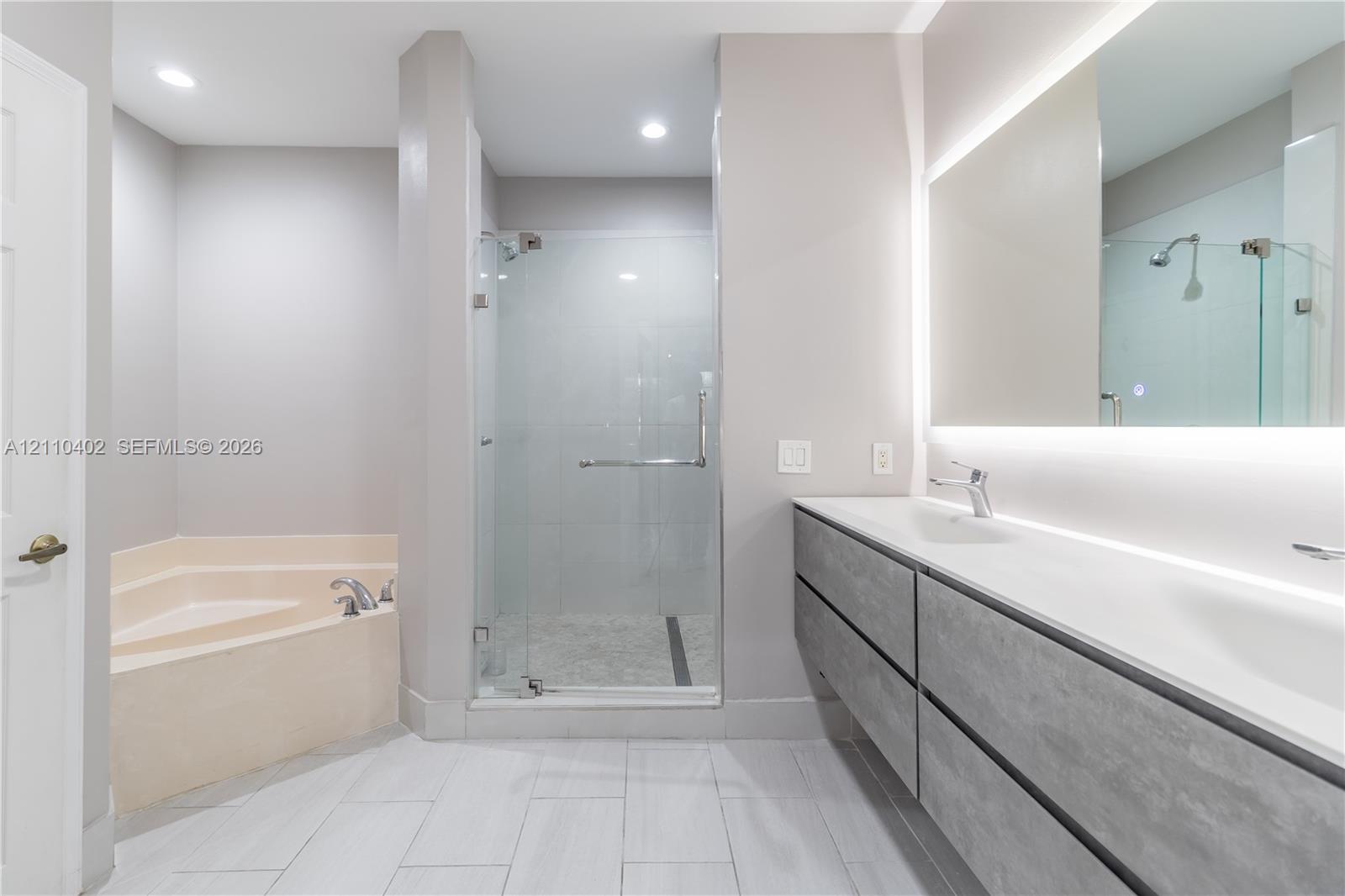 Floating vanity with stone-finish cabinetry, frameless glass-enclosed shower, large corner soaking tub, and illuminated vanity mirror