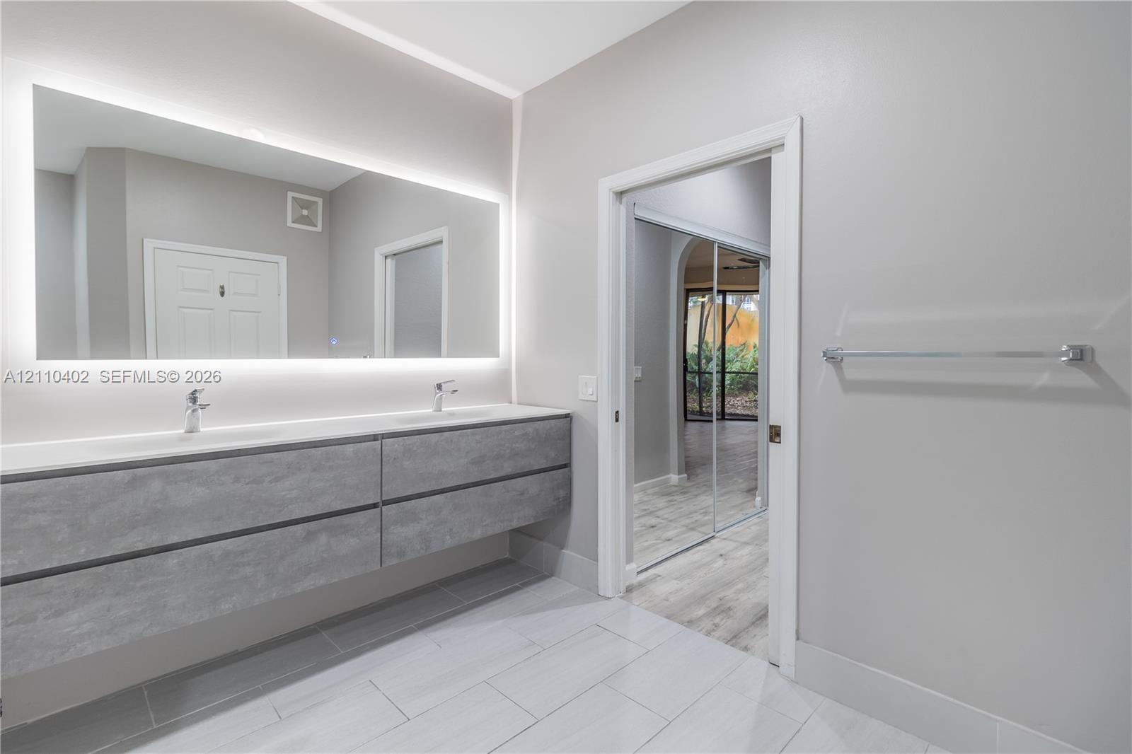 Floating vanity with a stone-finish facade, dual integrated sinks, and polished chrome faucets, complemented by a wall-to-wall backlit mirror and large-format tile flooring