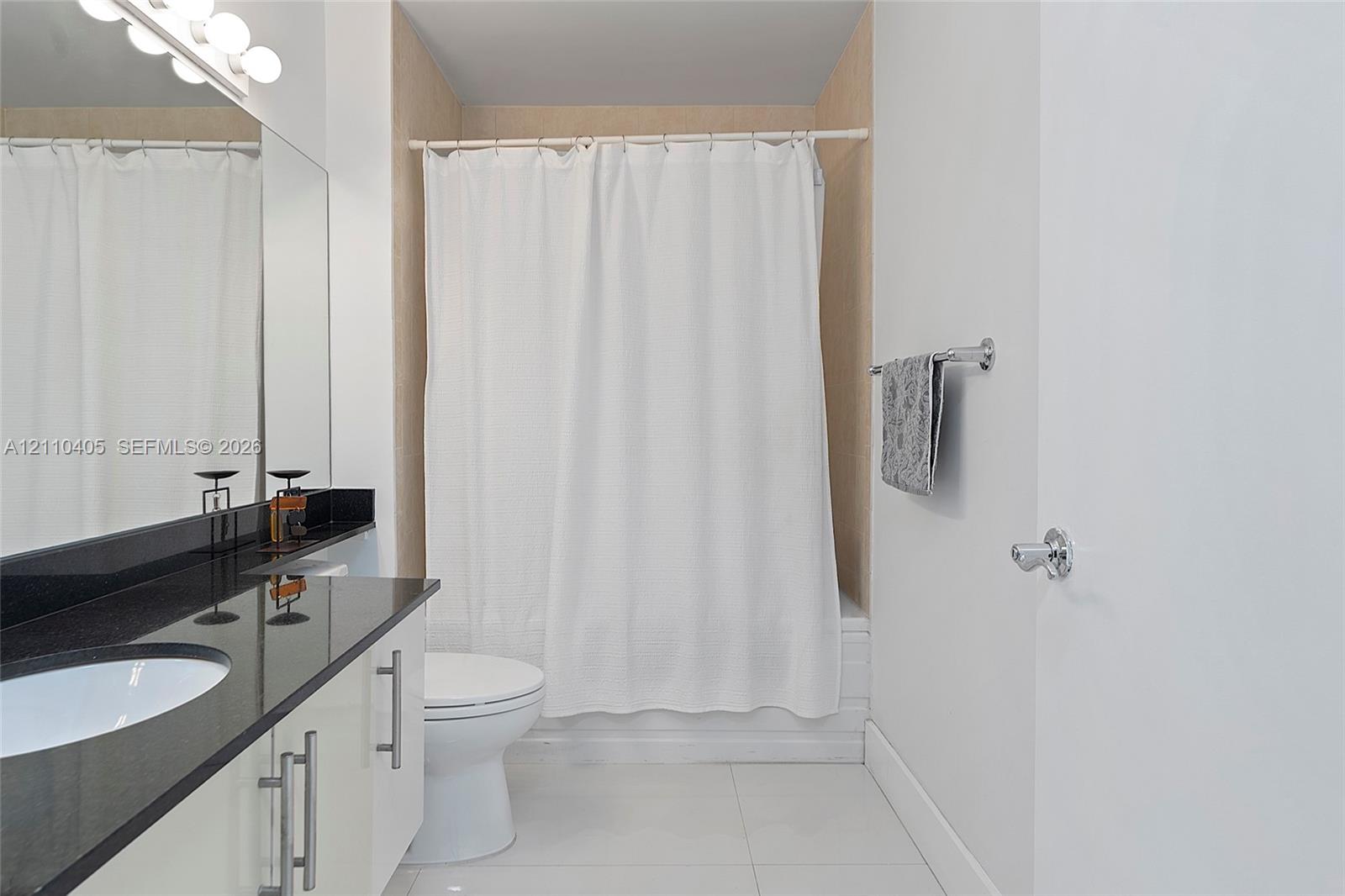 Bathroom featuring a vanity with dark stone countertop, integrated sink, polished metal hardware, full-width mirror with integrated lighting, and a bathtub with tile surround