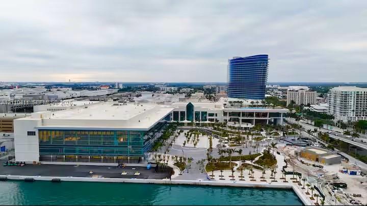 Aerial perspective of urban complex featuring waterfront proximity, glass-curtain wall architecture, paved plaza areas, and mature palm landscaping
