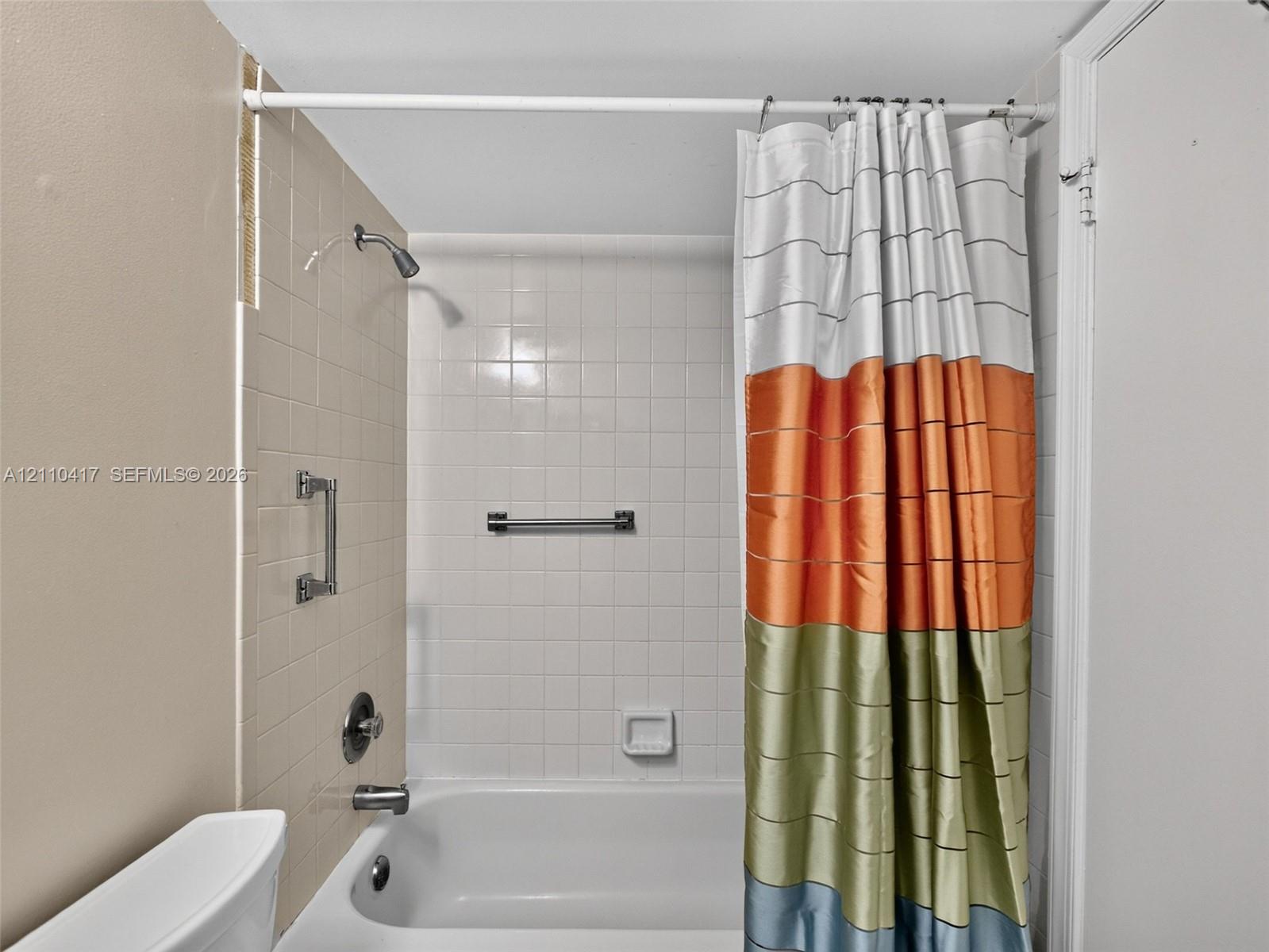 Bathtub and shower combination featuring a porcelain finish, wall-mounted shower head, integrated grab bar, and neutral square wall tiling