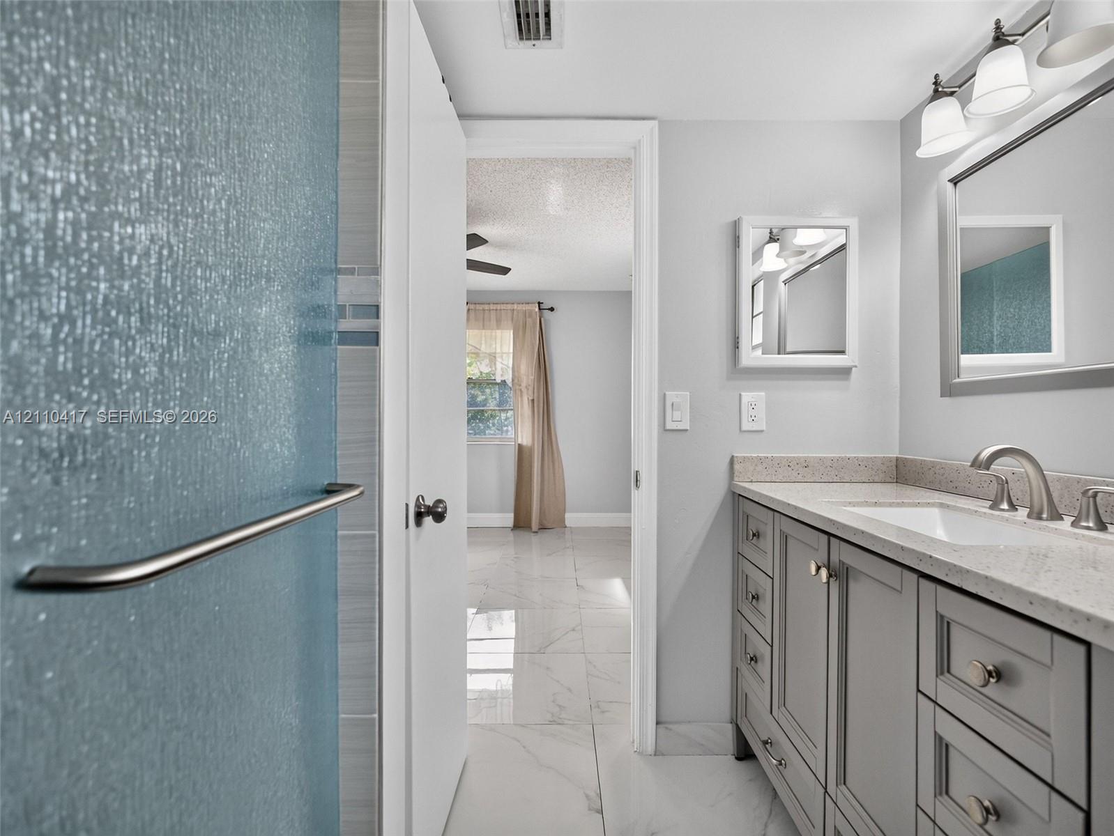 Bathroom featuring a light-toned vanity with stone-surface countertop, rectangular undermount sink, three-light wall fixture, and marble-finish flooring