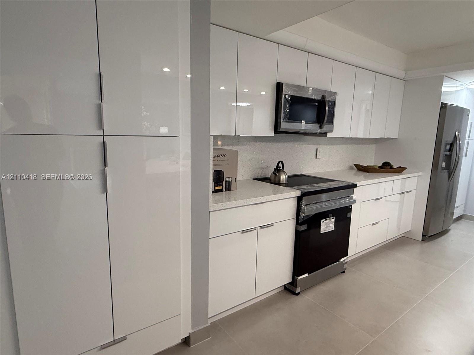 Kitchen featuring high-gloss white cabinetry, quartz countertops, tile backsplash, built-in stainless steel microwave, and large-format floor tile