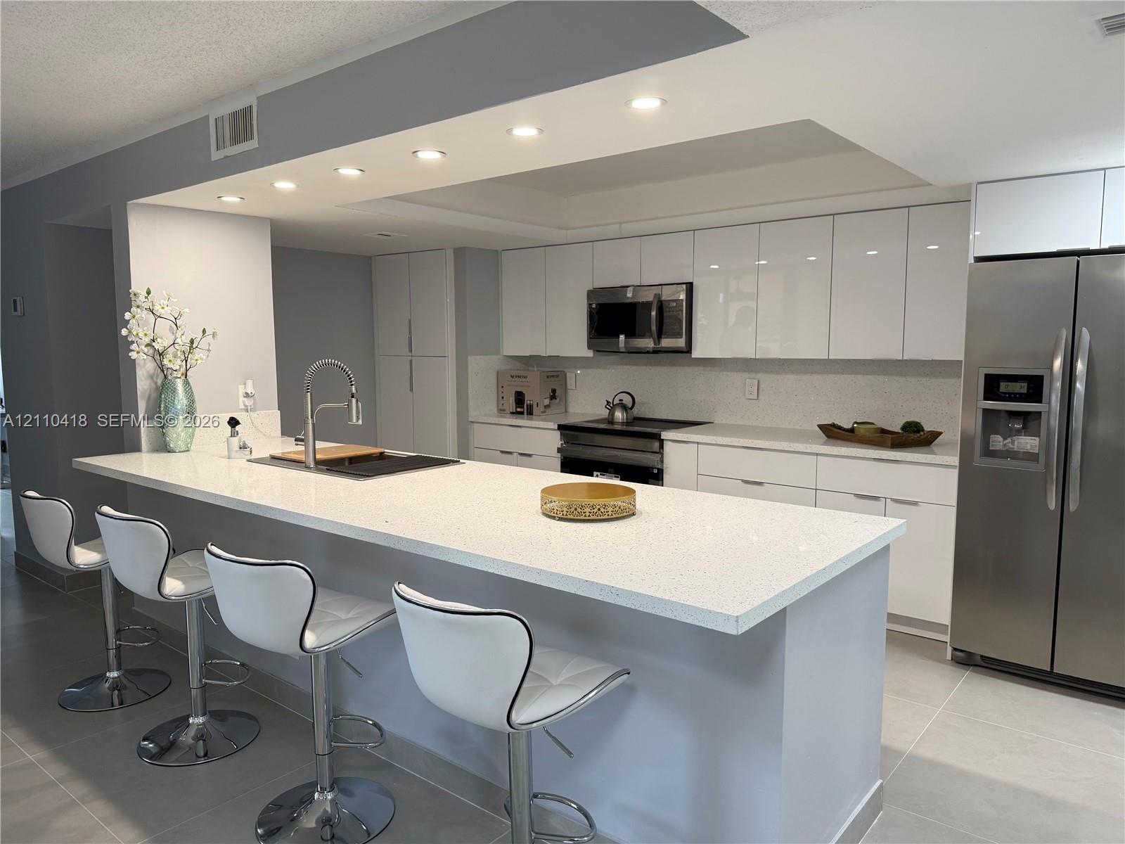 Kitchen featuring a large waterfall-edge island, high-gloss white cabinetry, solid-surface countertops, recessed ceiling lighting, and stainless steel appliances