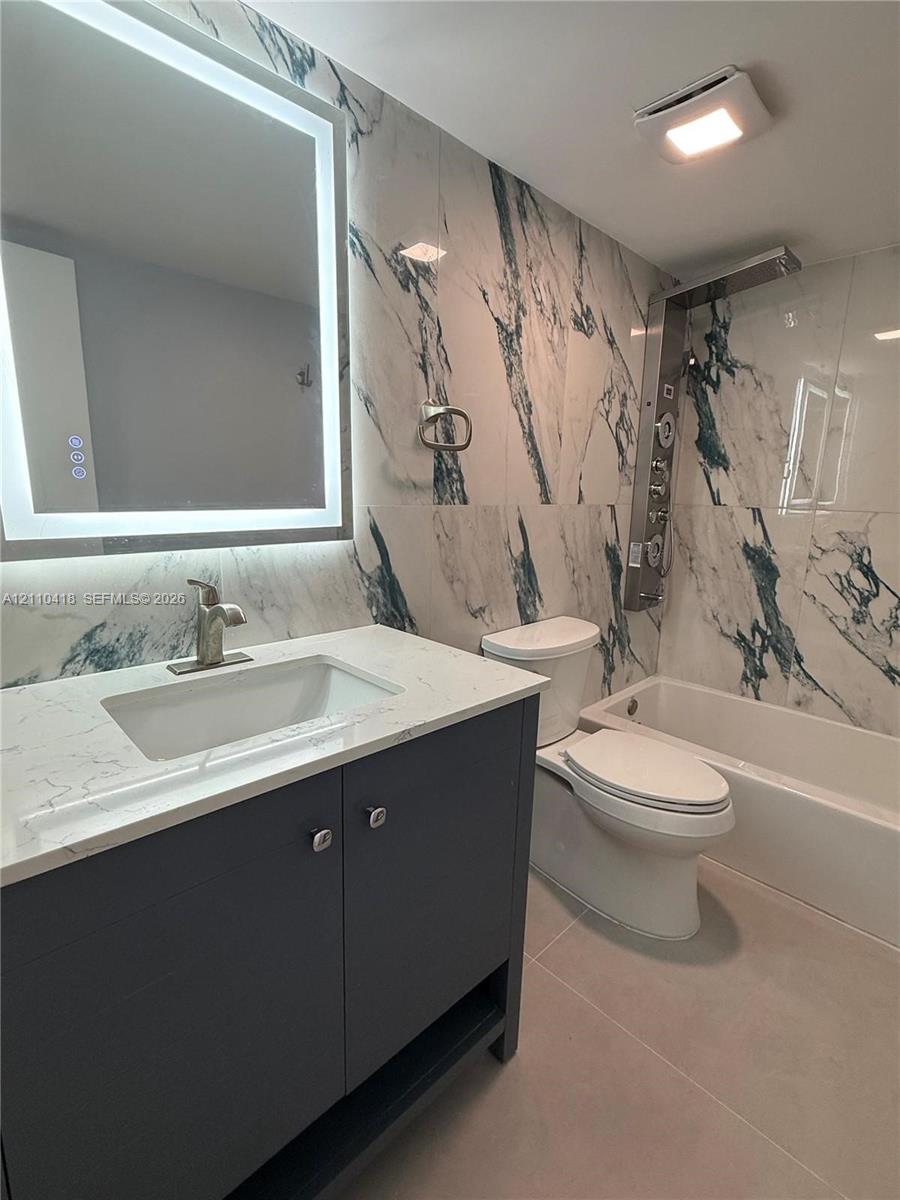 Full-bathroom featuring a dark finish vanity with a rectangular sink, quartz-surfaced countertop, LED-backlit mirror, large-format patterned wall tile, and a multi-head shower panel system