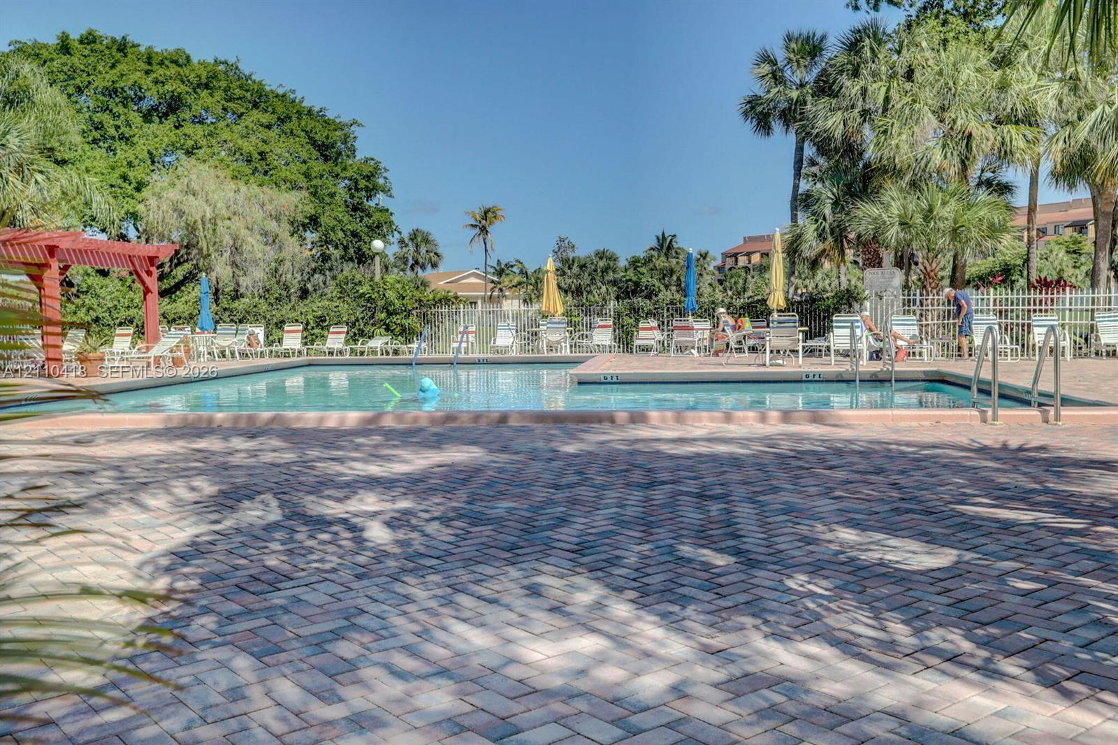 Outdoor swimming pool featuring a brick paver deck, perimeter metal fencing, and a red wood-finish pergola