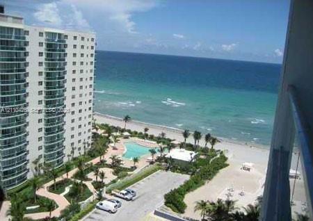 High-rise building with glass-paneled balconies overlooking a coastal landscape featuring a swimming pool, paved walking paths, landscaped grounds, and direct oceanfront access High-rise building with glass-paneled balconies overlooking a coastal landscape featuring a swimming pool, paved walking paths, landscaped grounds, and direct oceanfront access