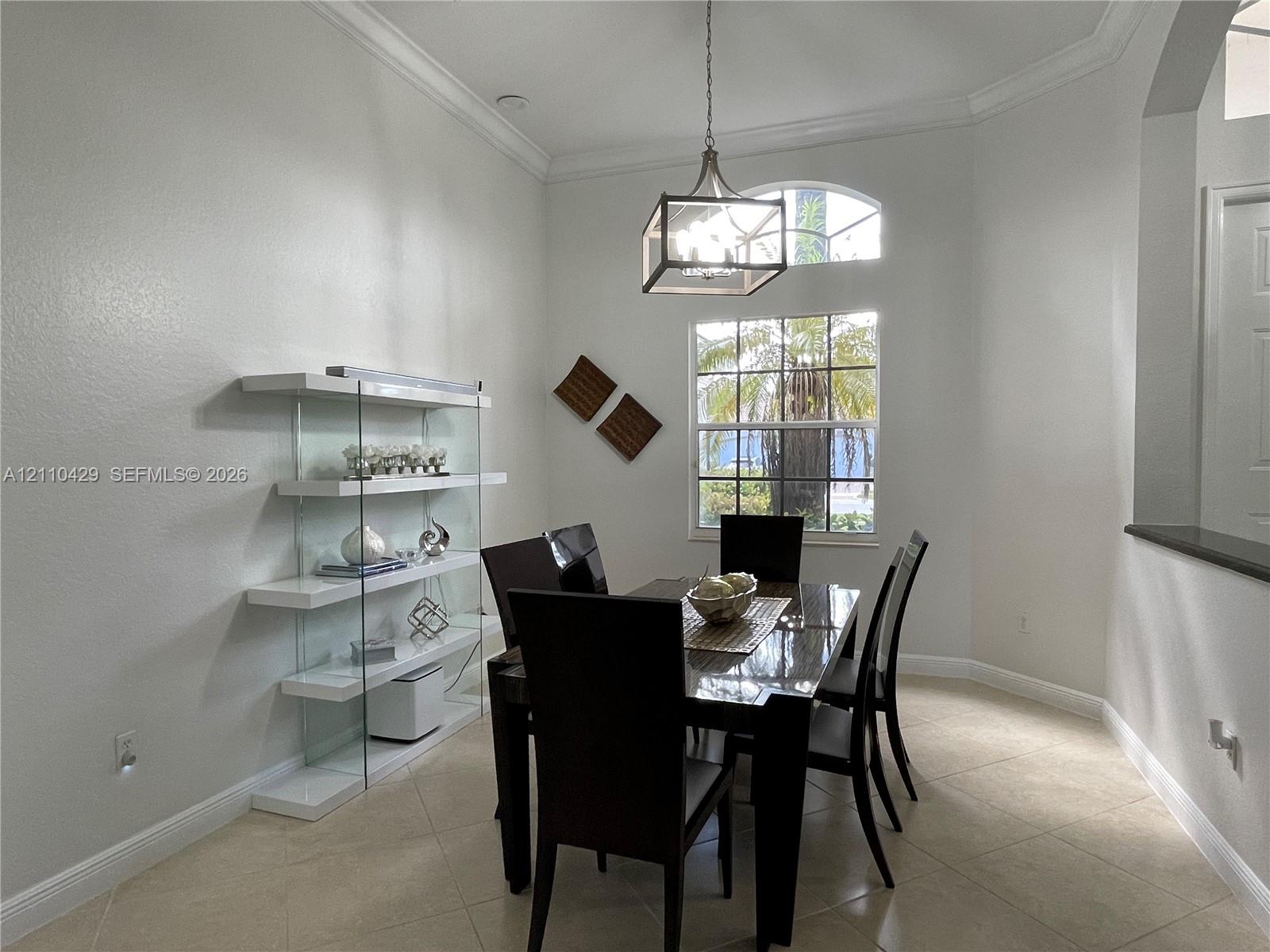 High-ceiling room featuring tile flooring, crown molding, a built-in glass and white shelving unit, and a metal-frame pendant light fixture