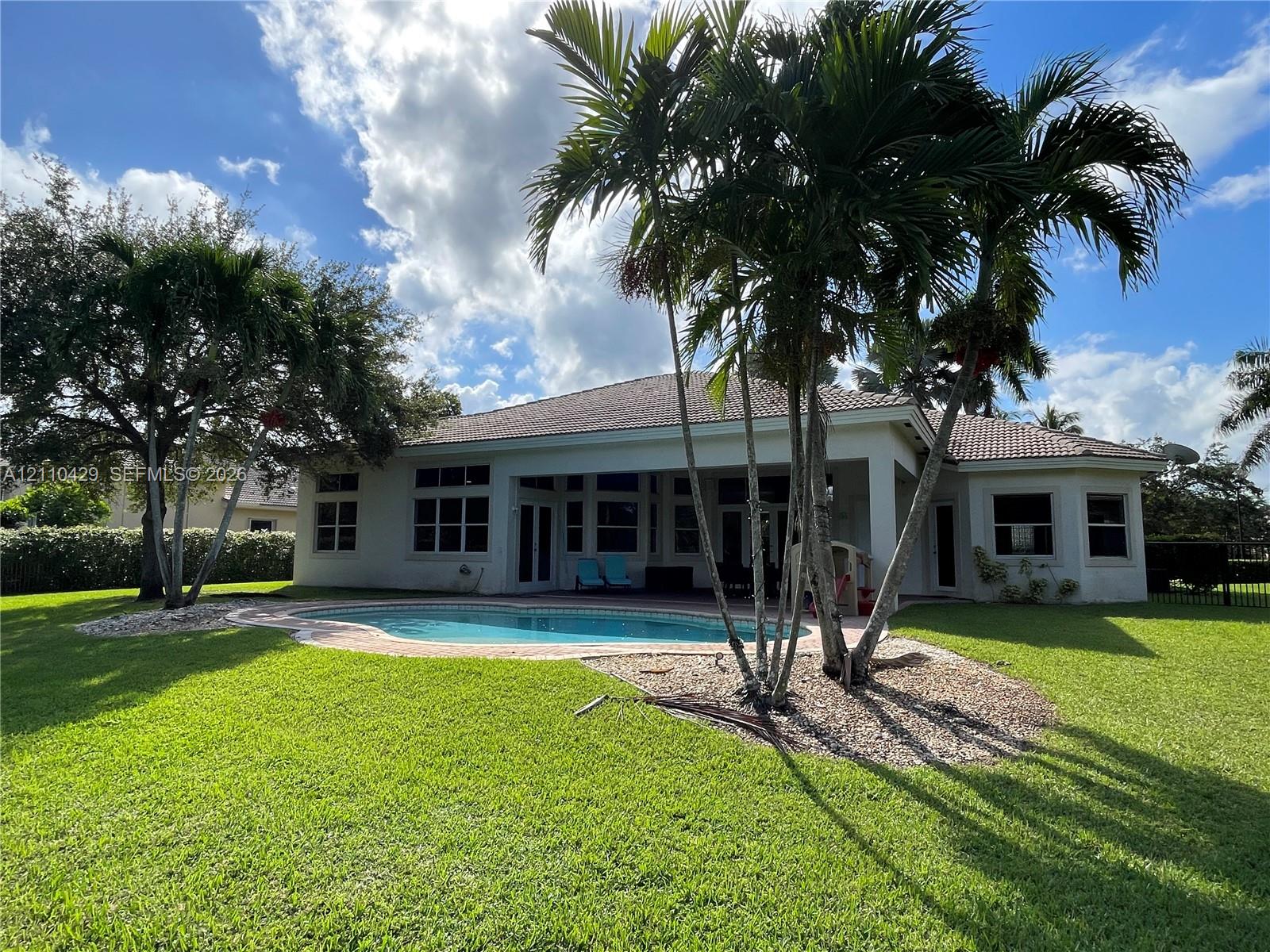Rear exterior featuring a covered patio, freeform swimming pool, manicured lawn, and mature palm trees