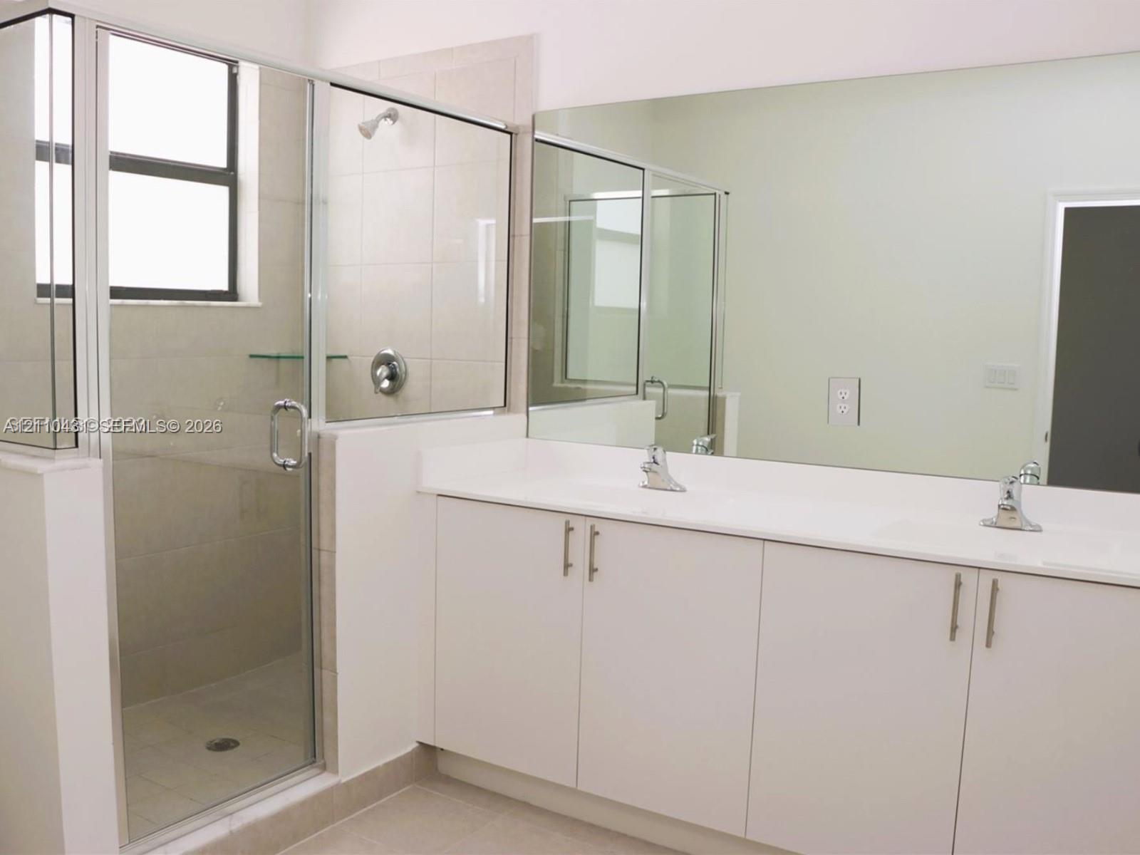 Double vanity with white cabinetry, solid-surface countertop, dual integrated sinks, and polished chrome faucets, complemented by a walk-in shower with glass enclosure and full-width mirror