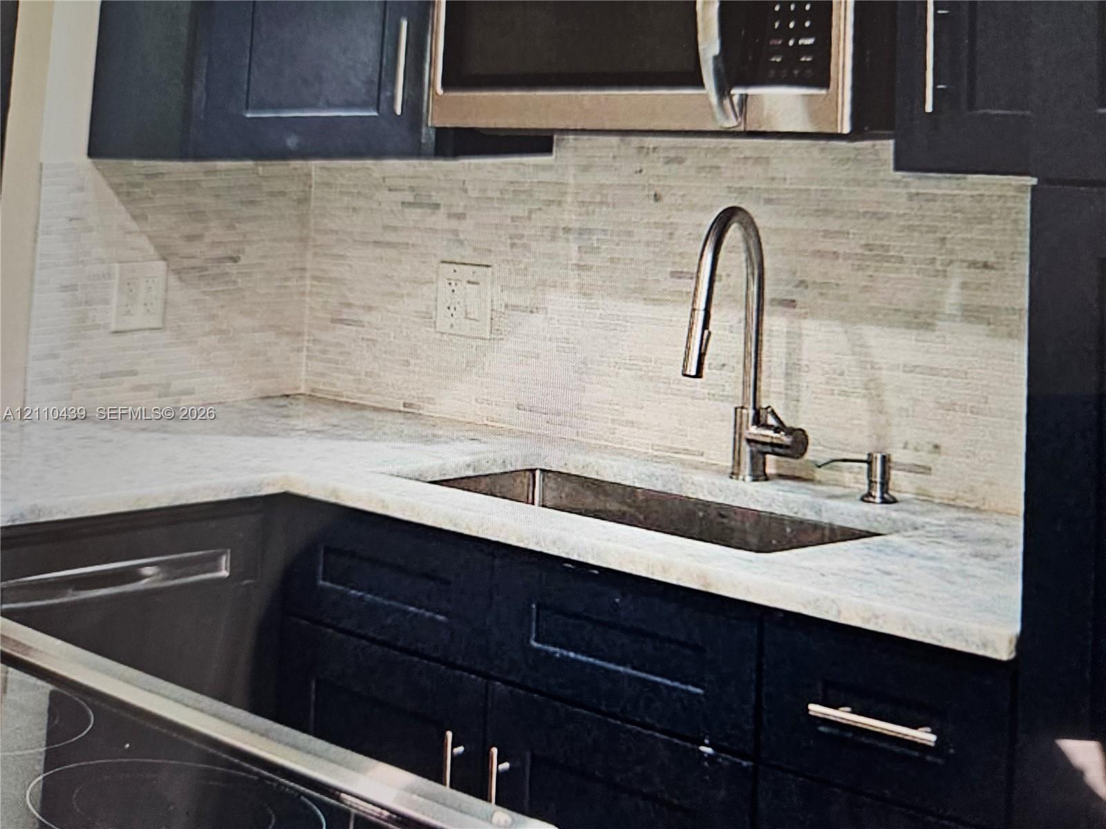 Kitchen featuring dark-toned cabinetry, light-toned stone-finish countertops, linear tile backsplash, stainless steel under-mount sink, and gooseneck pull-down faucet