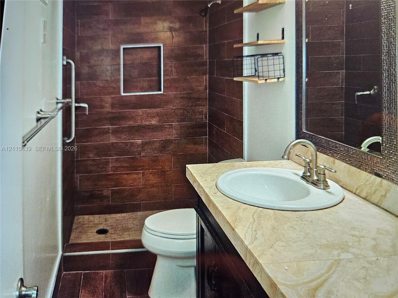 Walk-in shower featuring dark wood-finish tile walls and a built-in recessed niche, stone-finish vanity countertop with an undermount sink, and wall-mounted shelving