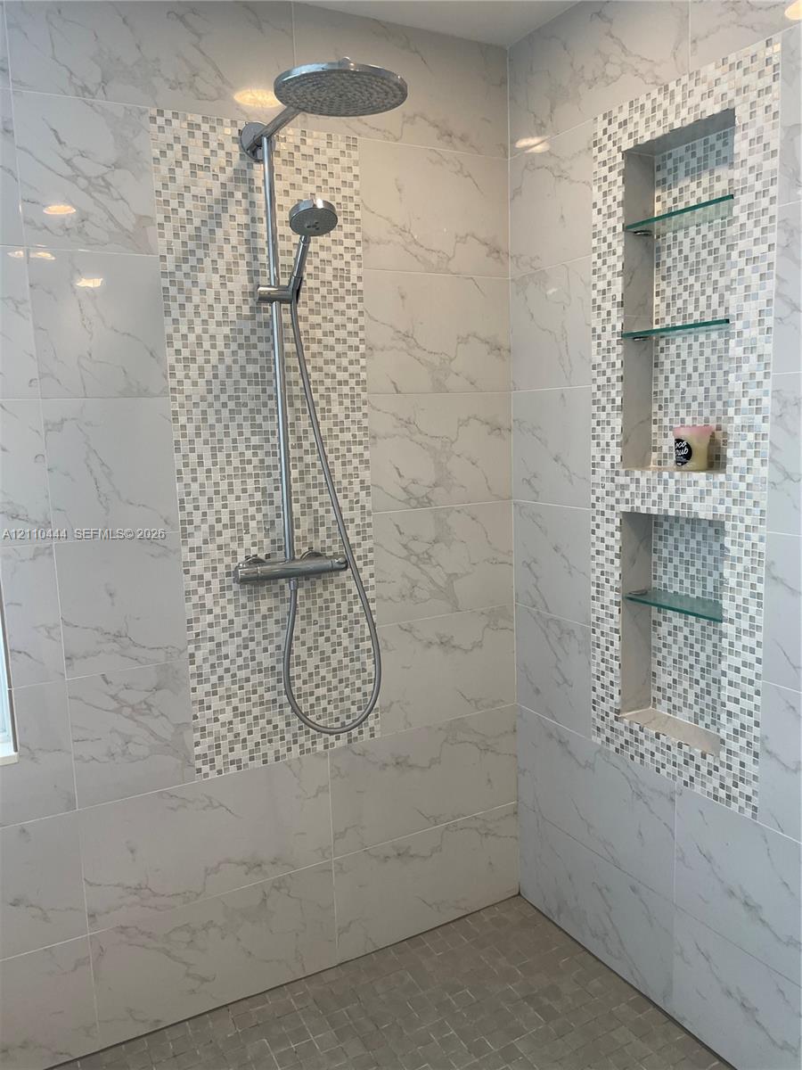 Walk-in shower featuring large-format stone-patterned wall tiles, decorative mosaic accent tiling, a rainfall showerhead with a handheld sprayer, and a built-in recessed niche with glass shelving