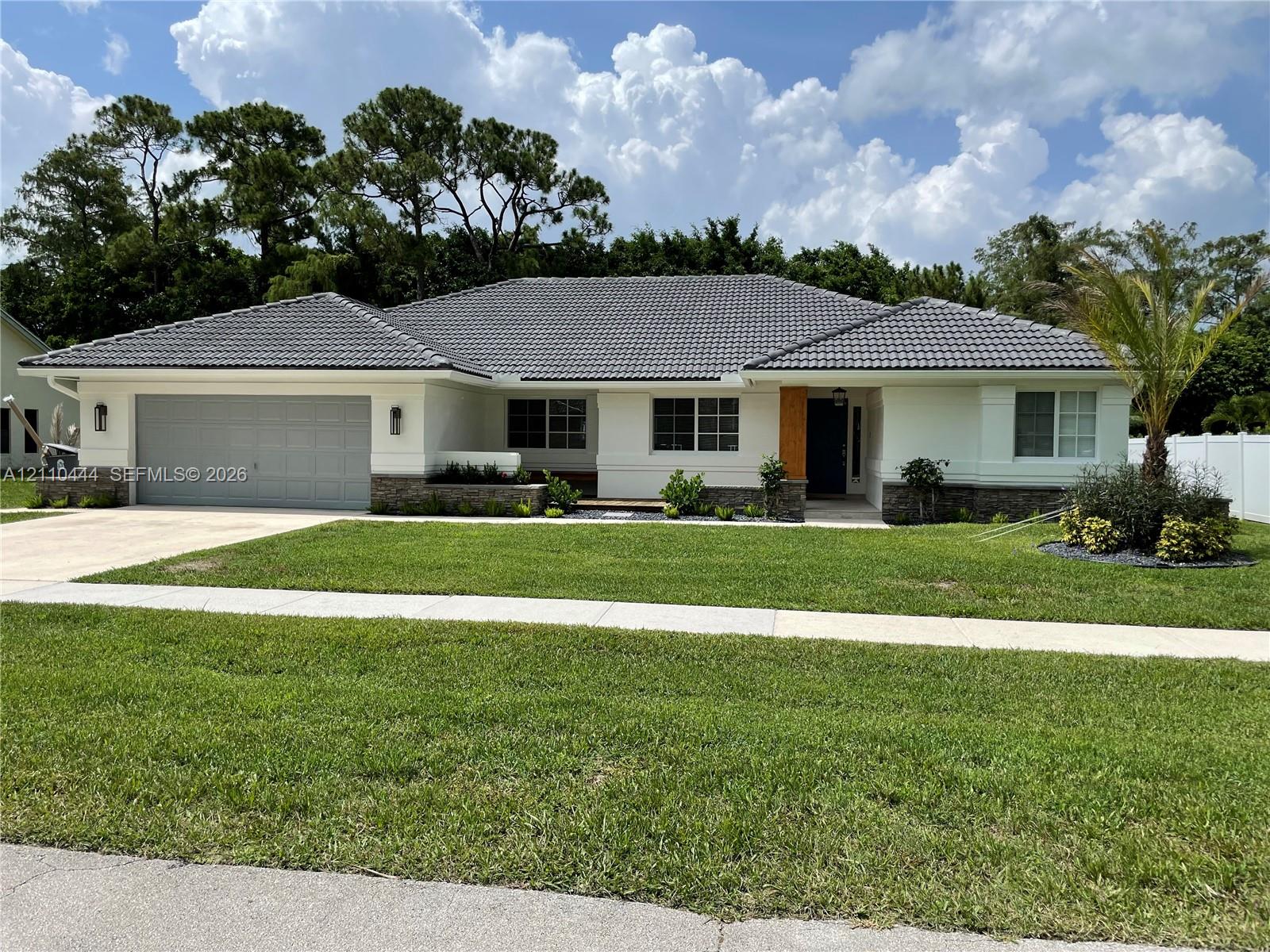 Single-story residence featuring a gray tiled roof, light-colored stucco exterior, stone masonry accents, a double-car garage, and a manicured lawn