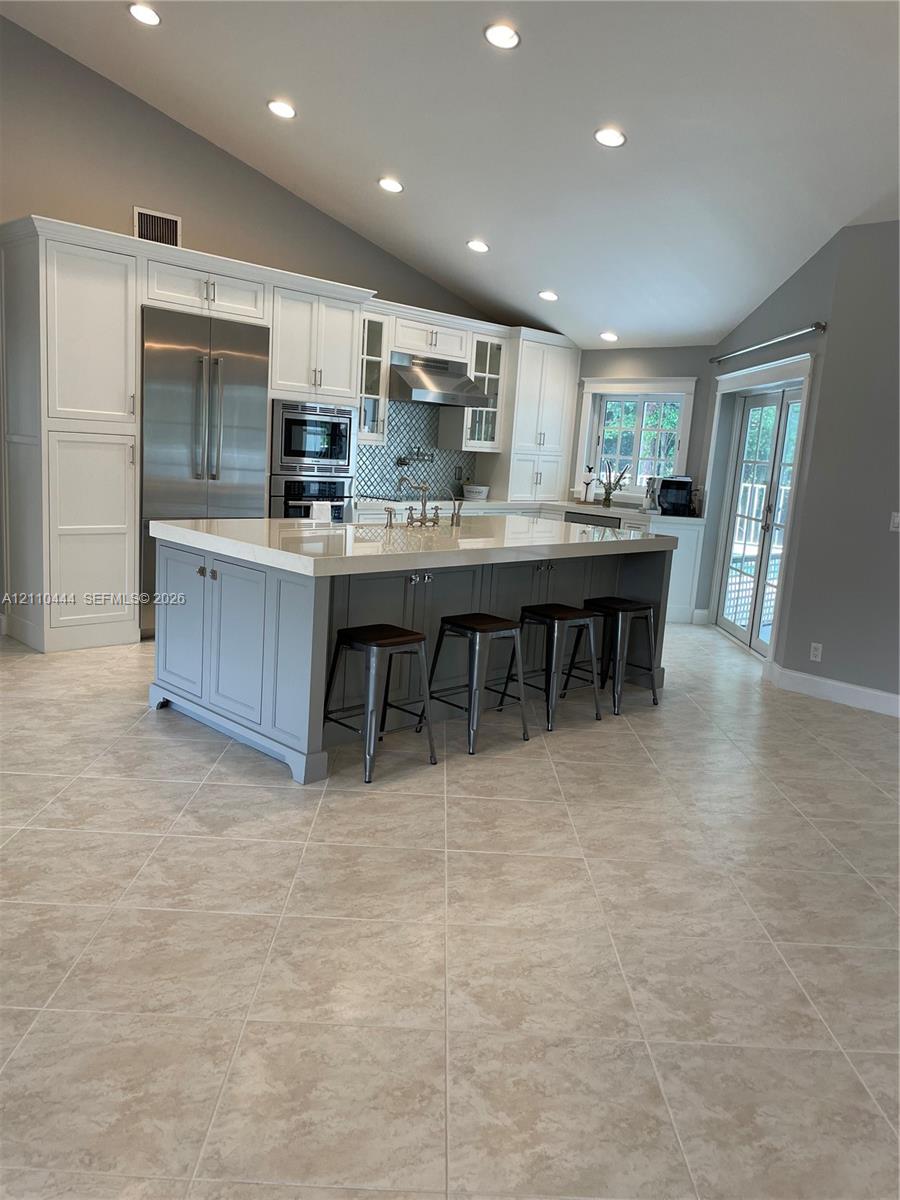 Kitchen featuring an oversized island with quartz countertops, custom cabinetry, stainless steel appliances, tile flooring, and vaulted ceilings with recessed lighting Kitchen featuring an oversized island with quartz countertops, custom cabinetry, stainless steel appliances, tile flooring, and vaulted ceilings with recessed lighting