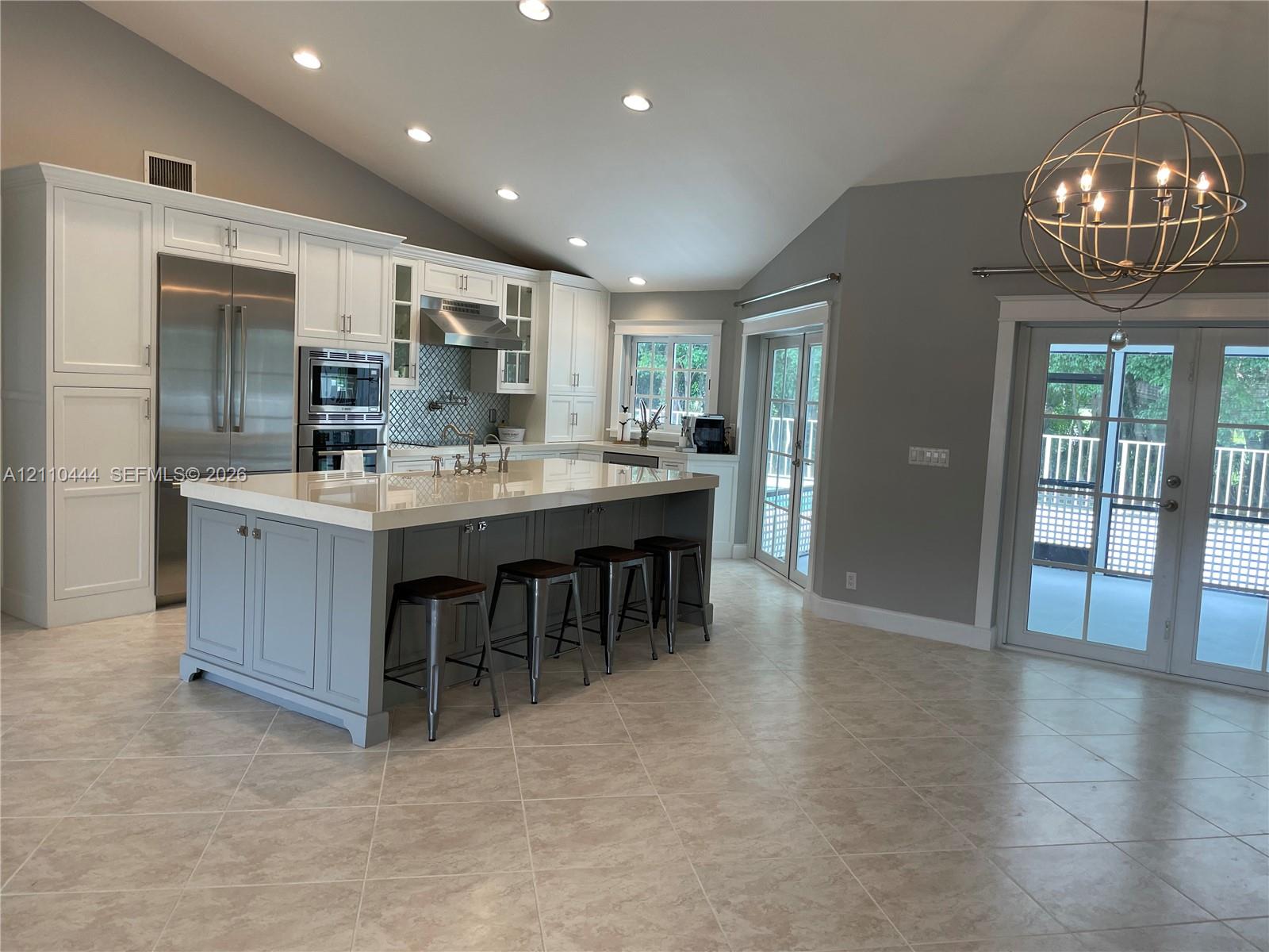 Kitchen featuring an oversized central island with stone-finish countertops, stainless steel appliances, vaulted ceilings, recessed lighting, and glass-paned doors Kitchen featuring an oversized central island with stone-finish countertops, stainless steel appliances, vaulted ceilings, recessed lighting, and glass-paned doors