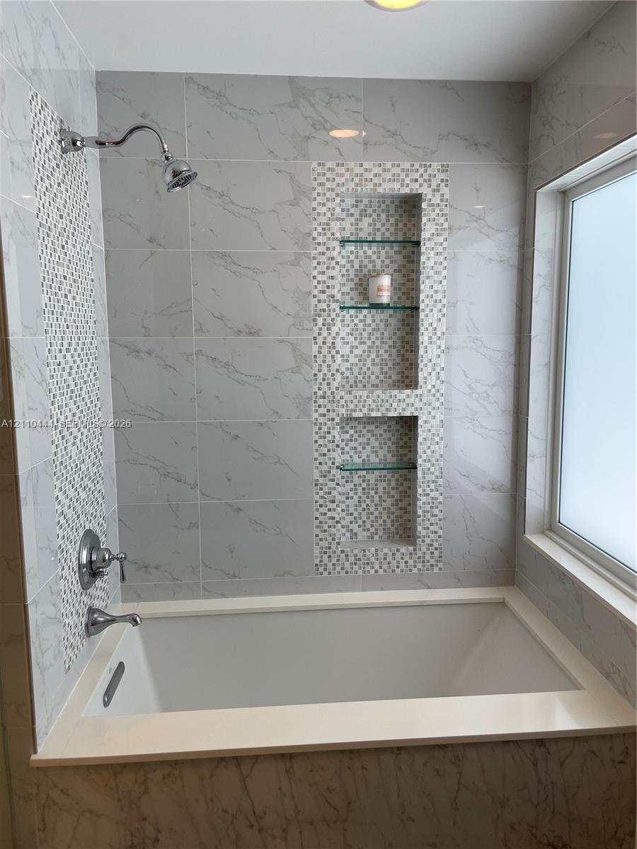 Bathtub surround featuring large-format veined tile, decorative mosaic tile accents, built-in recessed shelving with glass inserts, and polished chrome plumbing fixtures