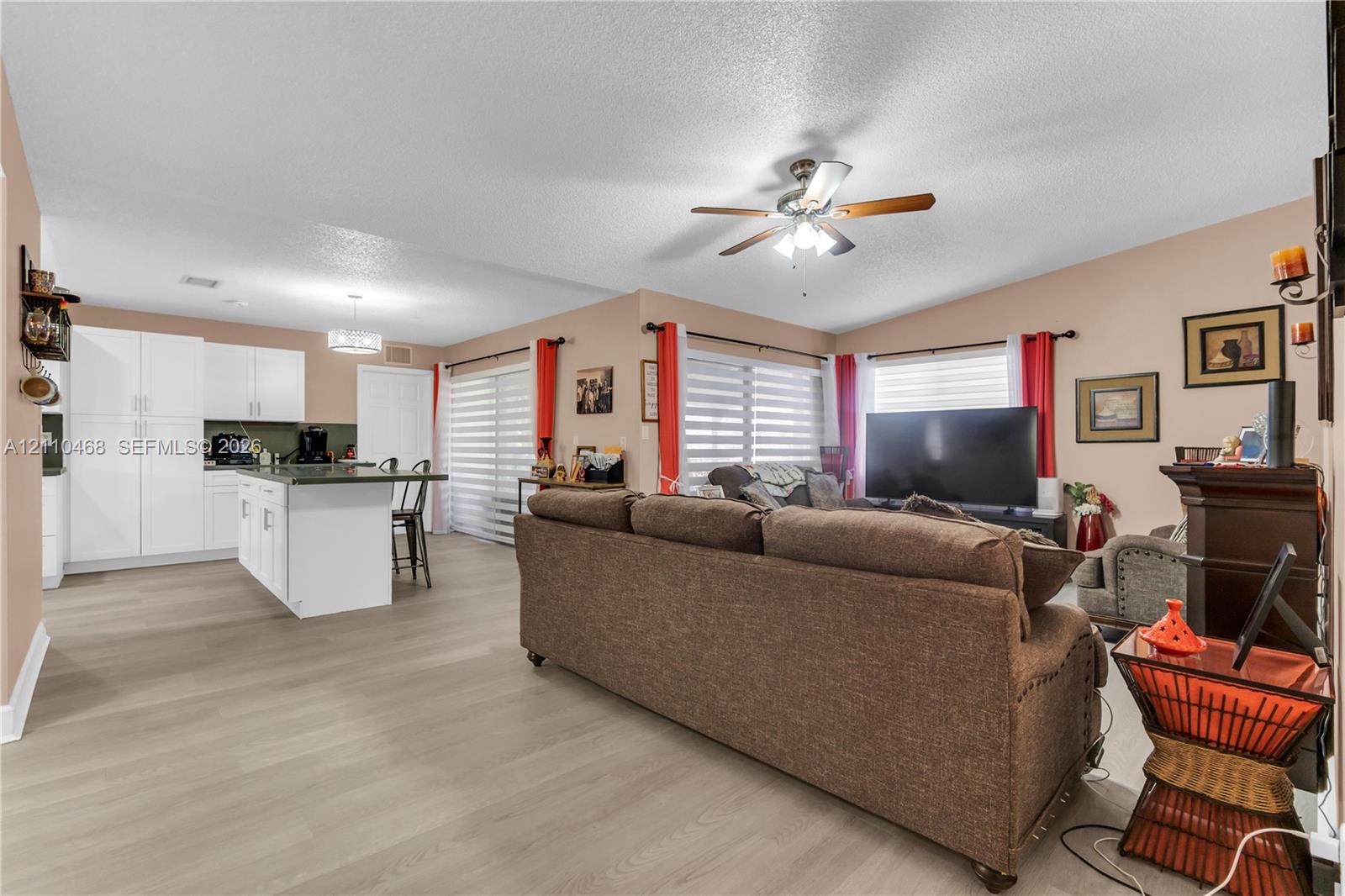 Open-concept interior featuring wood-finish flooring, a kitchen island, white cabinetry, and a ceiling fan with integrated lighting Open-concept interior featuring wood-finish flooring, a kitchen island, white cabinetry, and a ceiling fan with integrated lighting