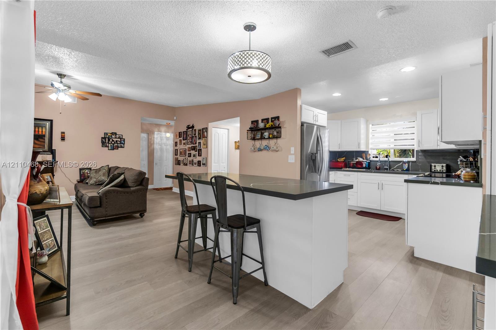 Open-concept kitchen featuring white cabinetry, dark countertops, a center island with breakfast bar seating, wood-finish flooring, and a modern drum pendant light fixture