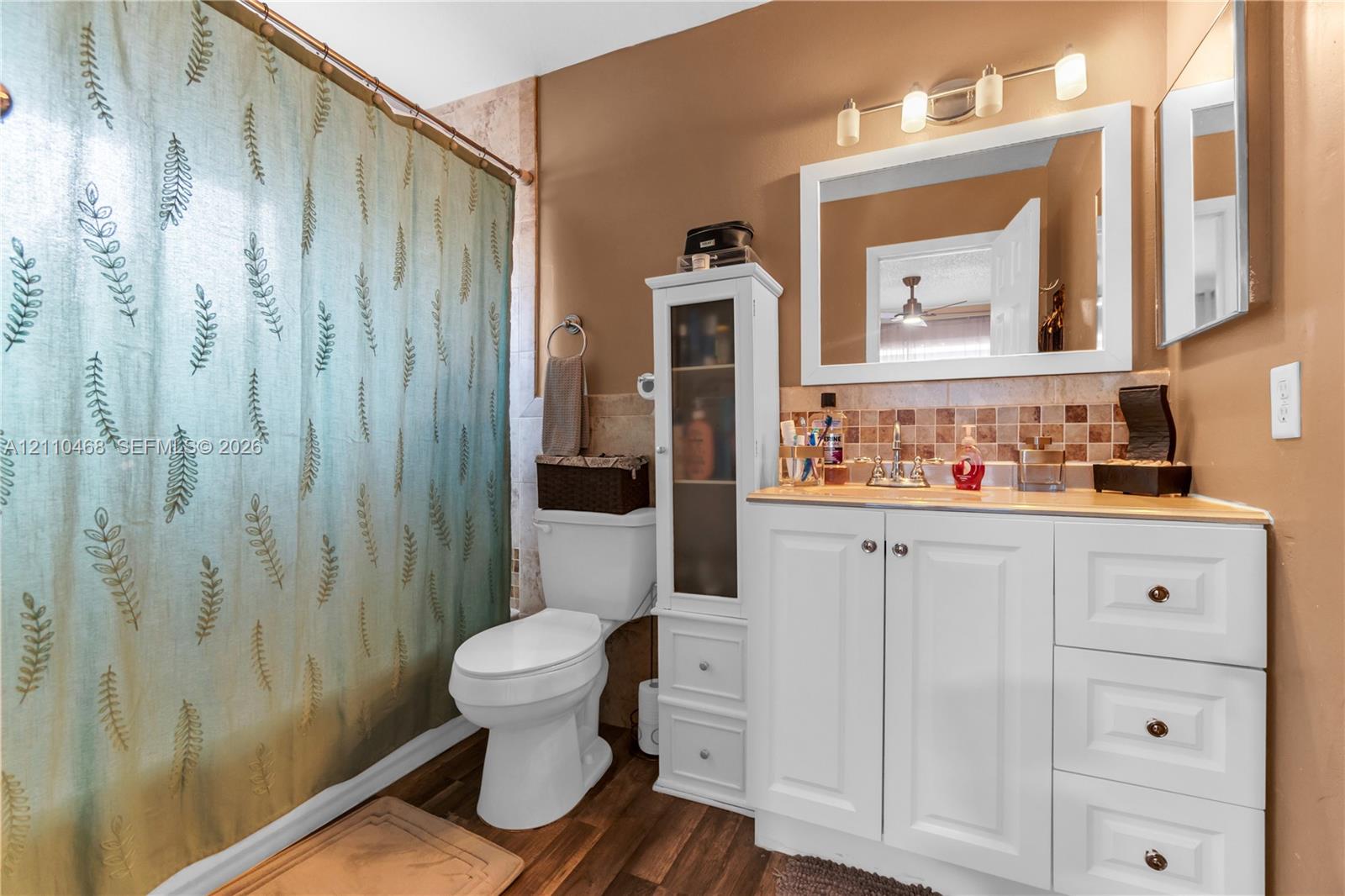 Bathroom featuring white vanity cabinetry with integrated storage tower, wood-finish flooring, multi-light vanity fixture, and tiled backsplash