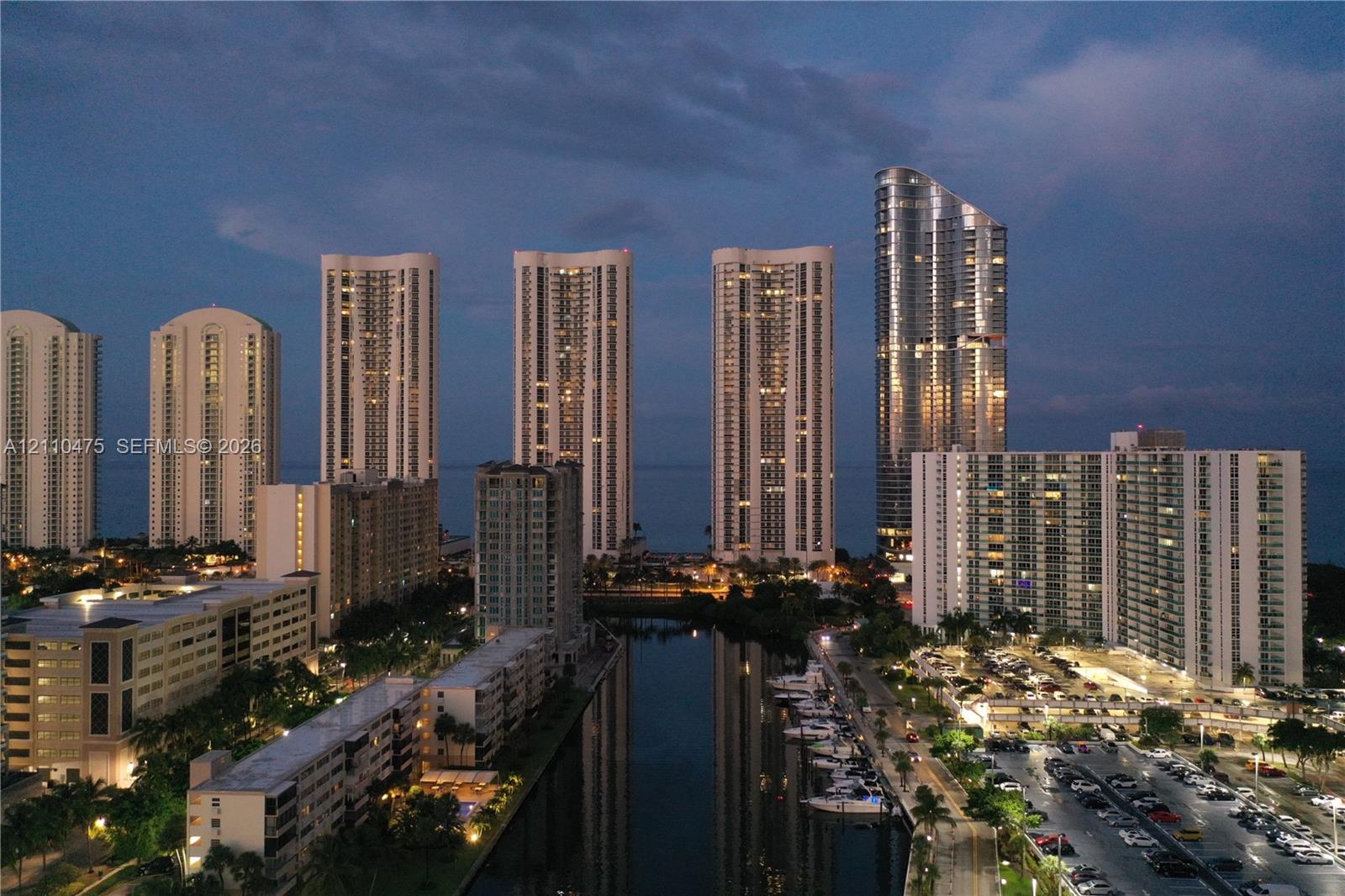 Aerial perspective of a coastal high-rise district featuring waterfront residential towers, a deep-water canal, paved parking facilities, and maritime docking slips