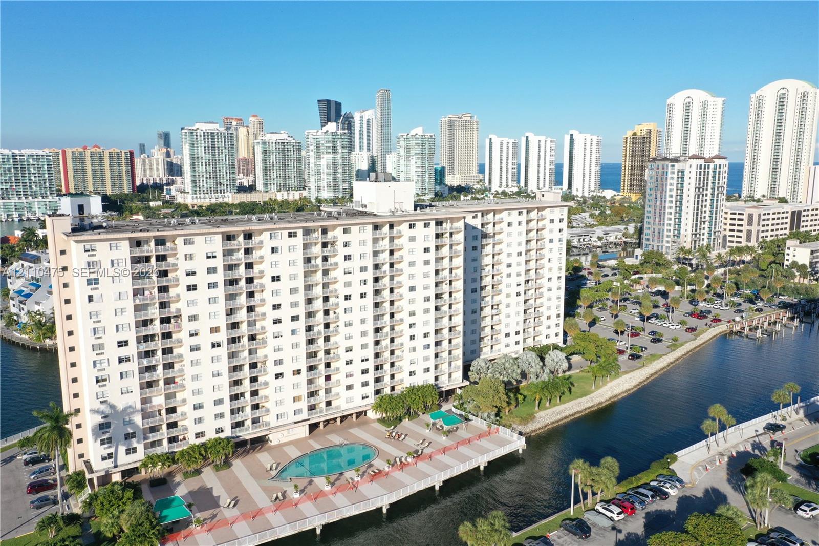 High-rise residential building featuring exterior balconies, a ground-level swimming pool, and concrete hardscaping situated along a waterway High-rise residential building featuring exterior balconies, a ground-level swimming pool, and concrete hardscaping situated along a waterway