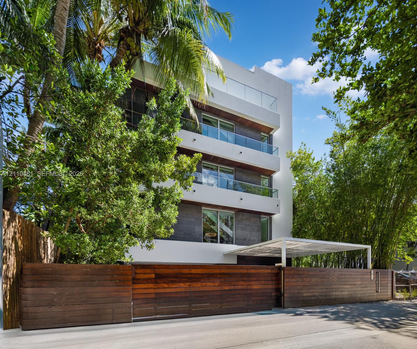 Modern multi-story exterior featuring white stucco and dark stone cladding, stacked balconies with glass railings, and a horizontal wood slat perimeter fence
